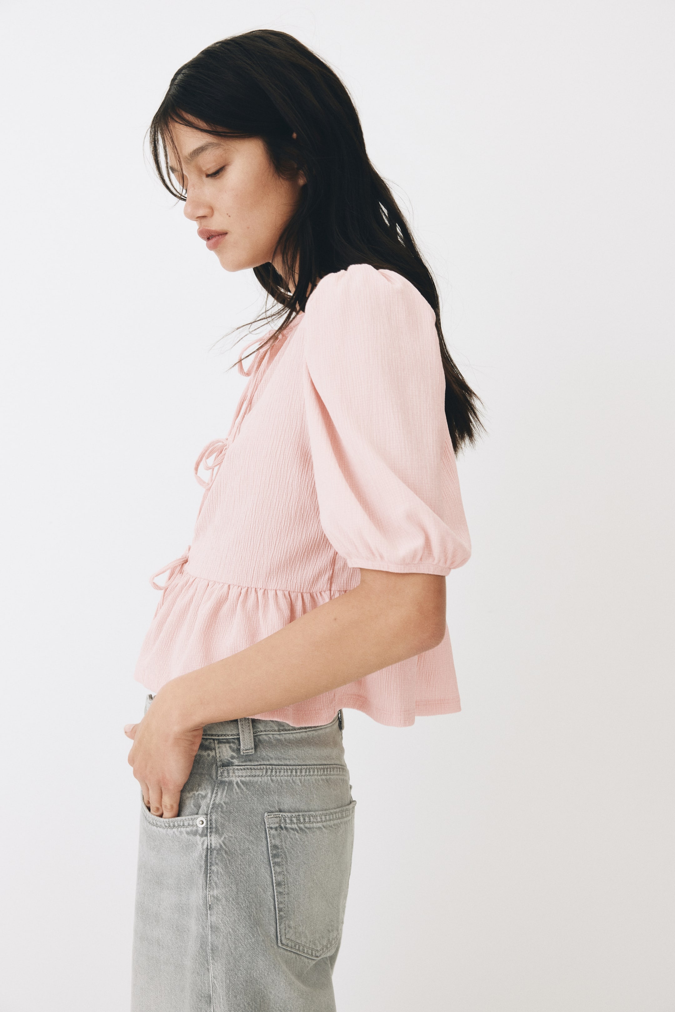 View larger image: The pink textured top showcases a tie-front closure, a gathered peplum hem, and puffed elbow-length sleeves with elasticated cuffs, presented from a side profile to highlight its fit and structure.