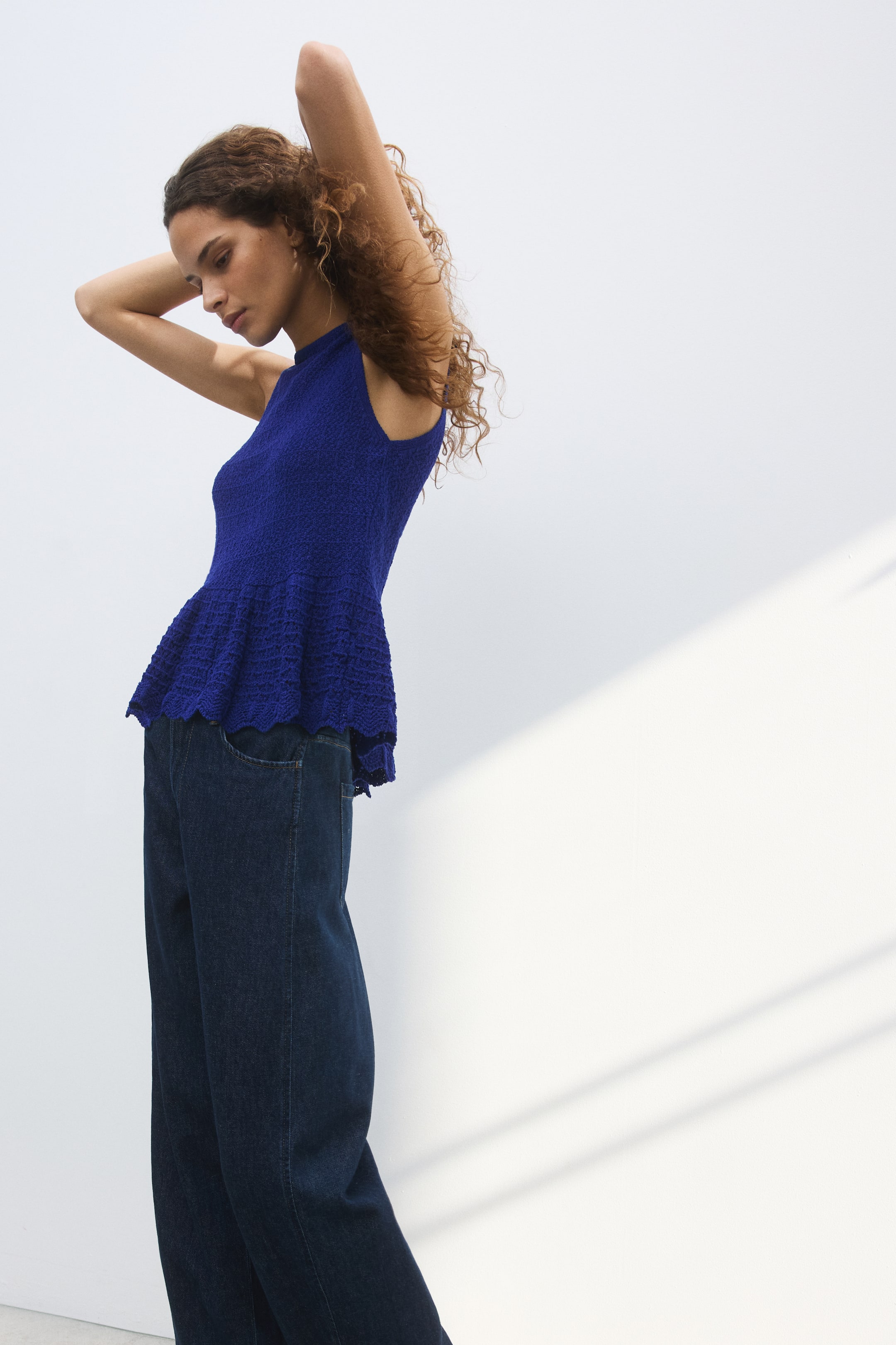 Se større billede: Den kongelige blå ærmeløse peplum-striktop har en høj halsudskæring og en tætsiddende overdel med en tæt, tekstureret strik. Dens flared peplum viser et åbent, mønstret strik og en bølget kant, der flyder over mørkeblå wide-leg jeans fra en sidevinkel.