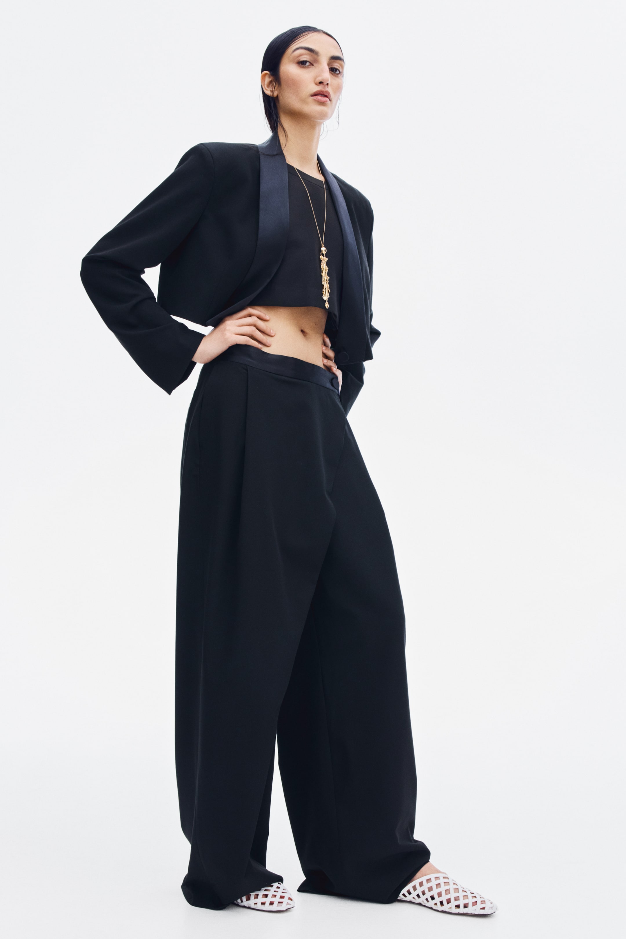 View larger image: Black wide-leg twill pants featuring a satin waistband, front pleats, and a wrap-front design are being worn by the model. The pants are styled with a coordinating cropped black blazer, a black crop top, and white woven shoes.