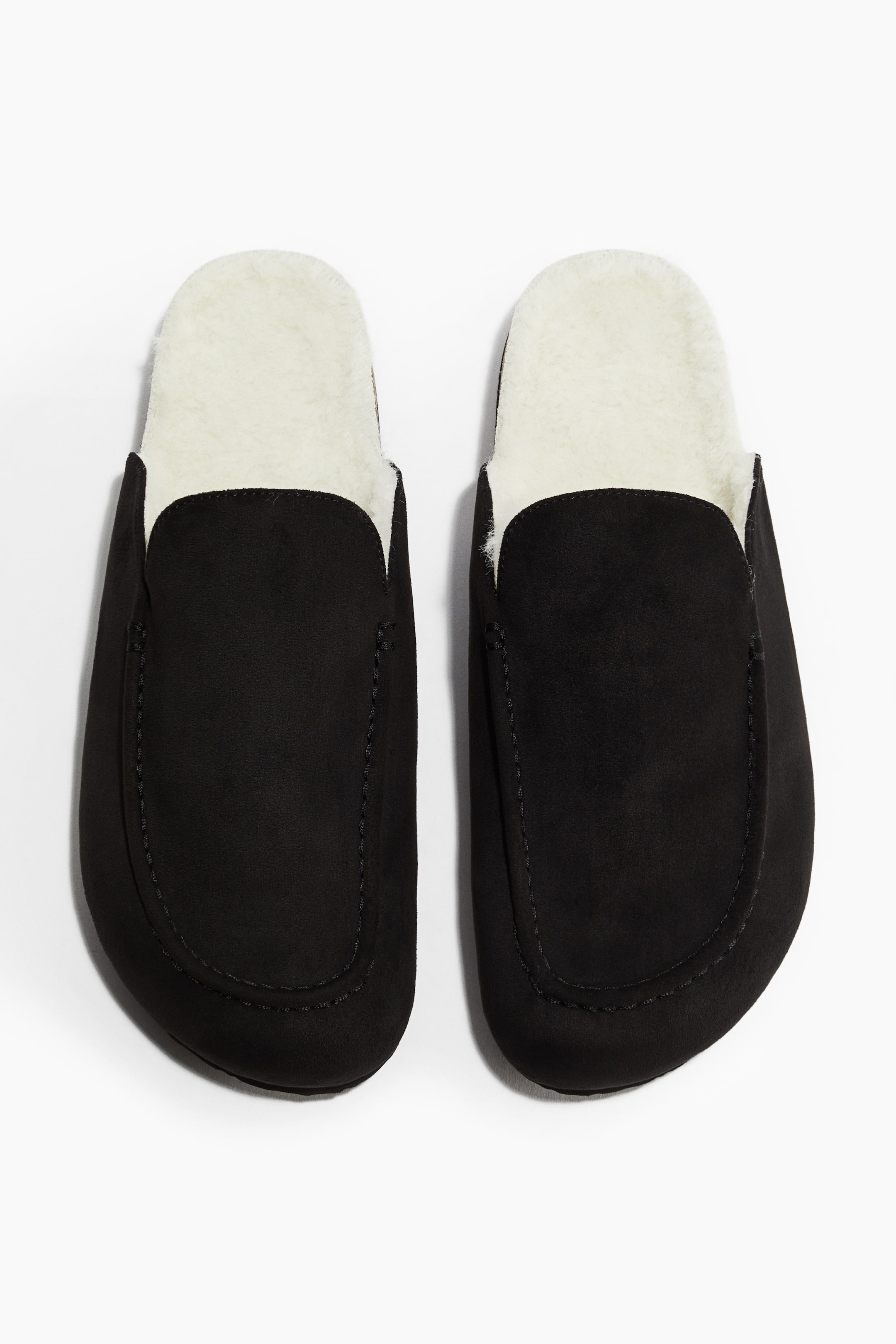 View larger image: A pair of black open-back slippers is being presented, showcasing a cream-colored fluffy lining that is extending around the opening and inside the footbed. Moccasin-style stitching is detailing the vamp of each slipper.