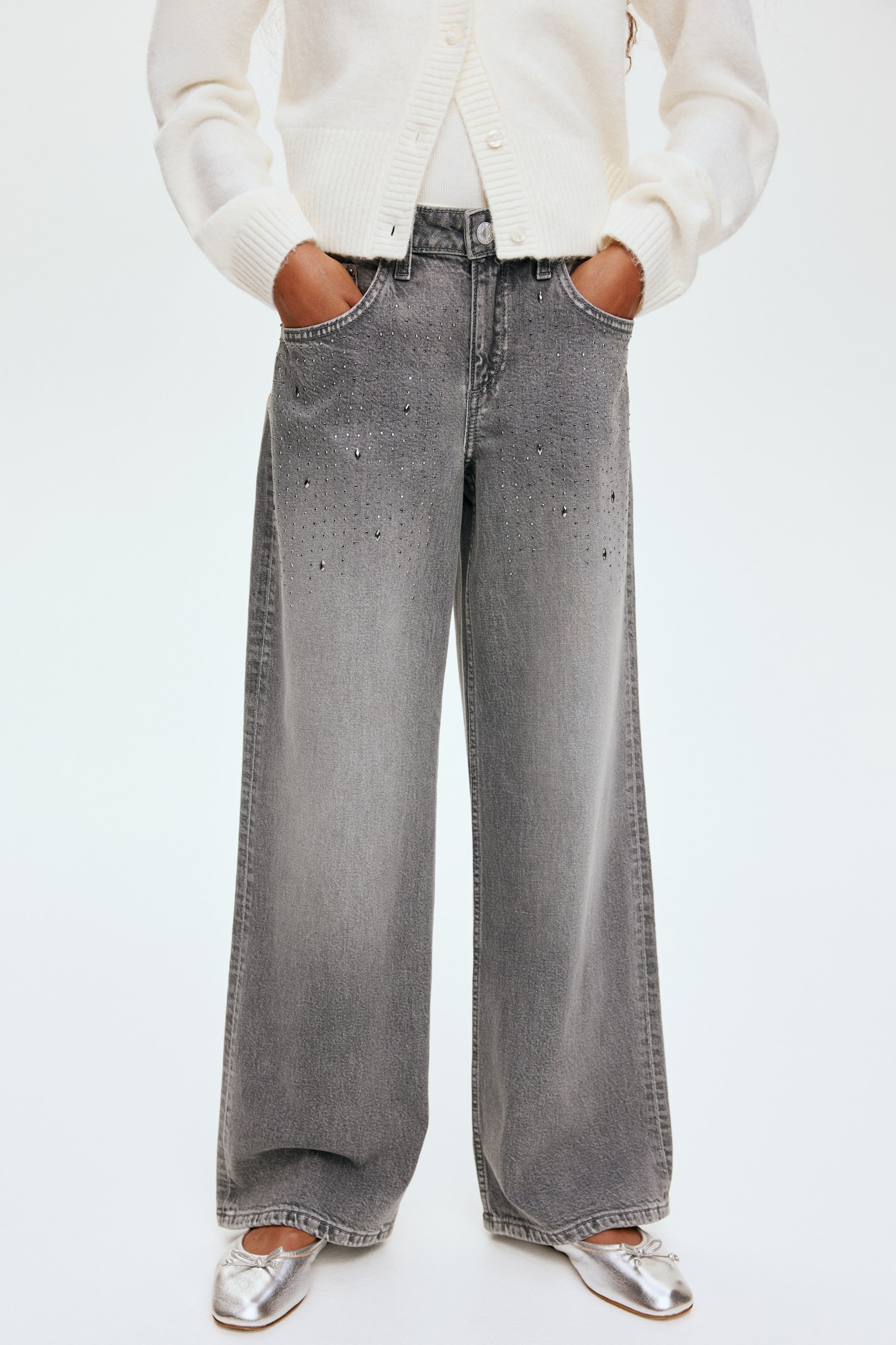 View larger image: Faded gray wide-leg jeans are being presented from the front, featuring scattered stud embellishments, more concentrated towards the upper thighs. The jeans are showcasing a loose fit with visible front pockets where hands are resting.
