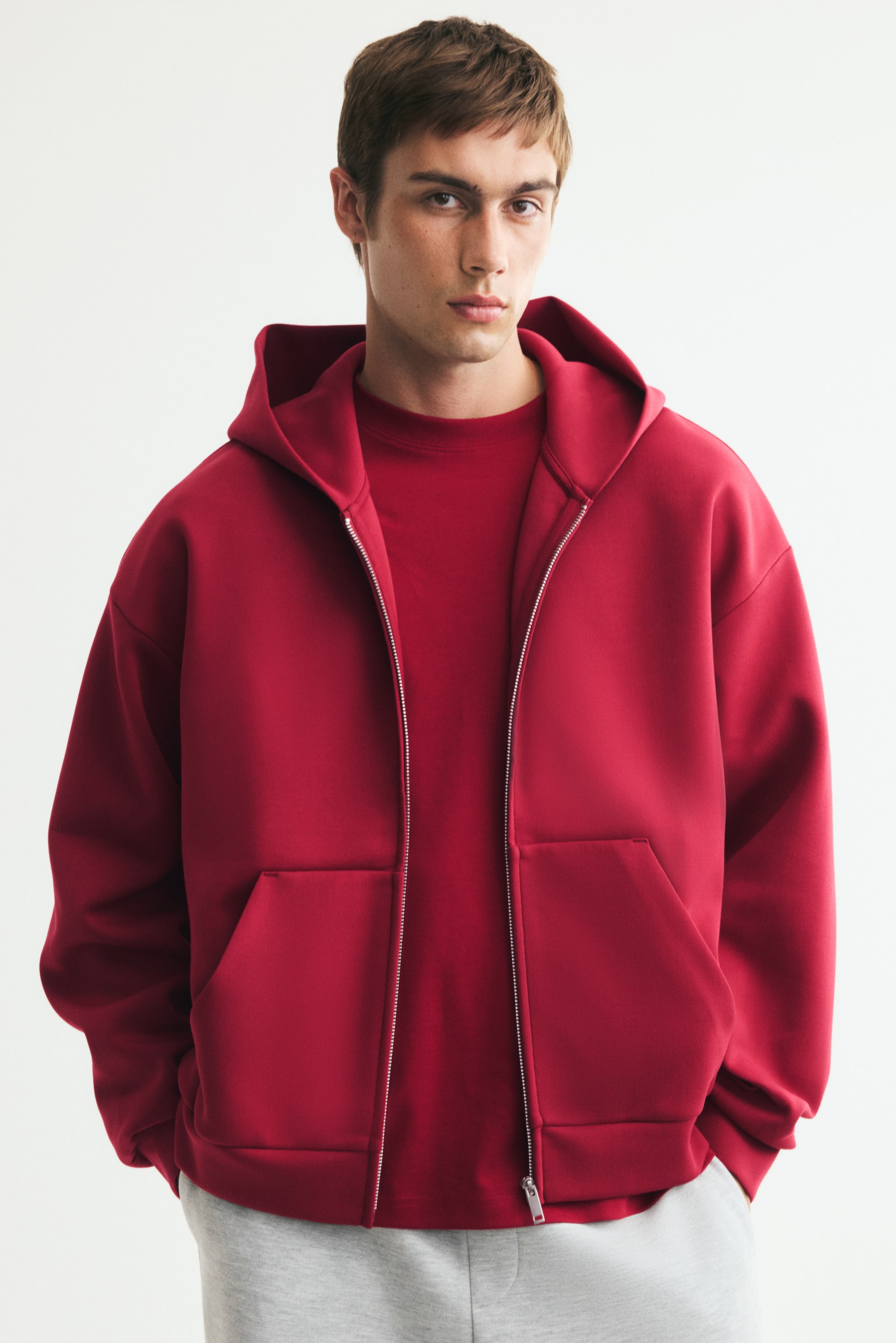 View larger image: A red zip-up hoodie in structured interlock jersey is being worn by the model over a matching red crewneck t-shirt and light gray pants. The hoodie features an oversized silhouette with dropped shoulders, front pockets, and a lined hood.