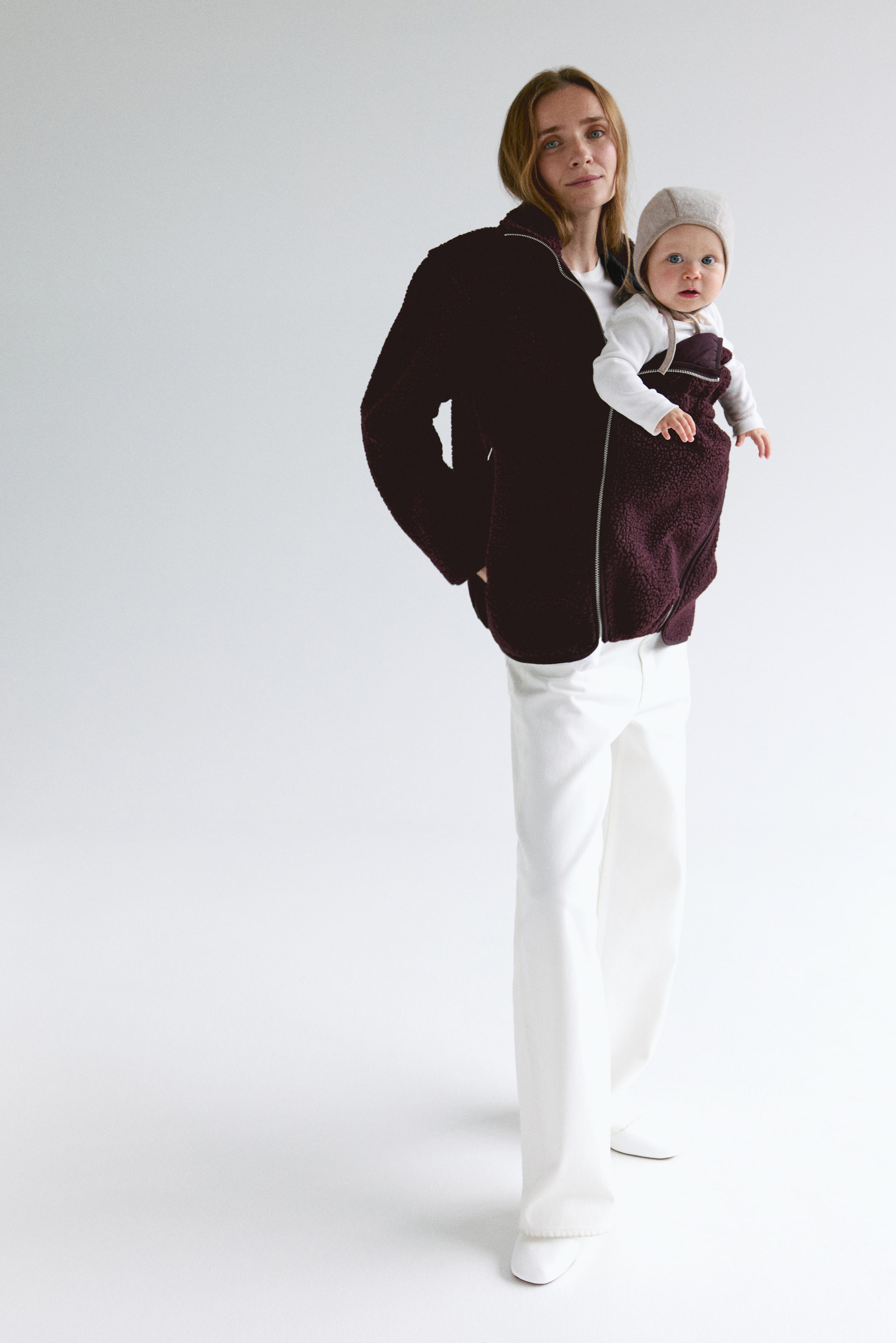 View larger image: The model is wearing a teddy jacket in a loose fit, with a stand collar, front zip, and zipped side pockets. The model is carrying a baby in the jacket’s detachable, jersey-lined baby panel, which features covered elastic at the top and bottom.