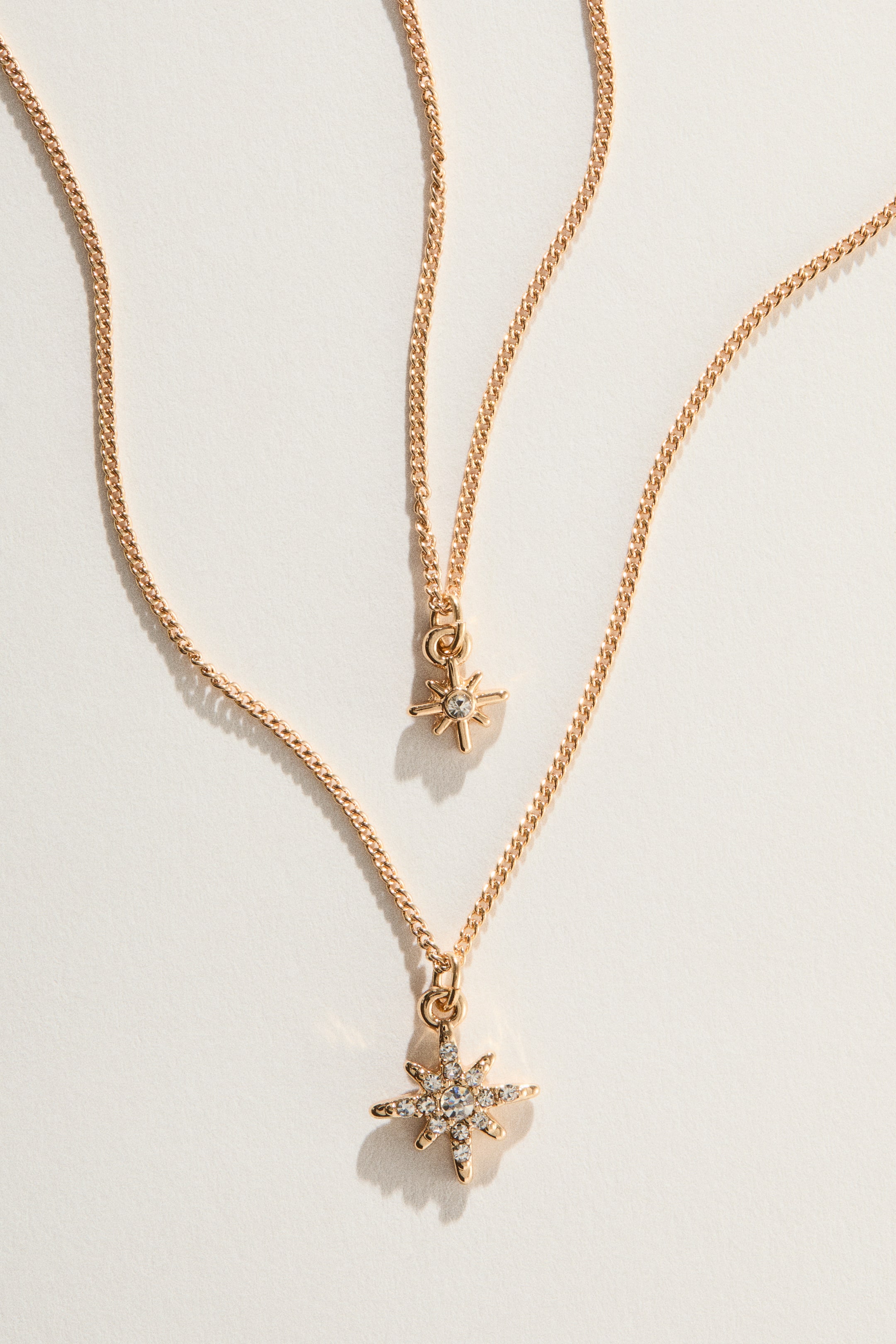 View larger image: A layered necklace features two gold-toned chains, each displaying a star-shaped pendant. The shorter chain’s star showcases a single clear stone at its centre, while the longer chain’s star is adorned with multiple clear stones surrounding a central clear stone.