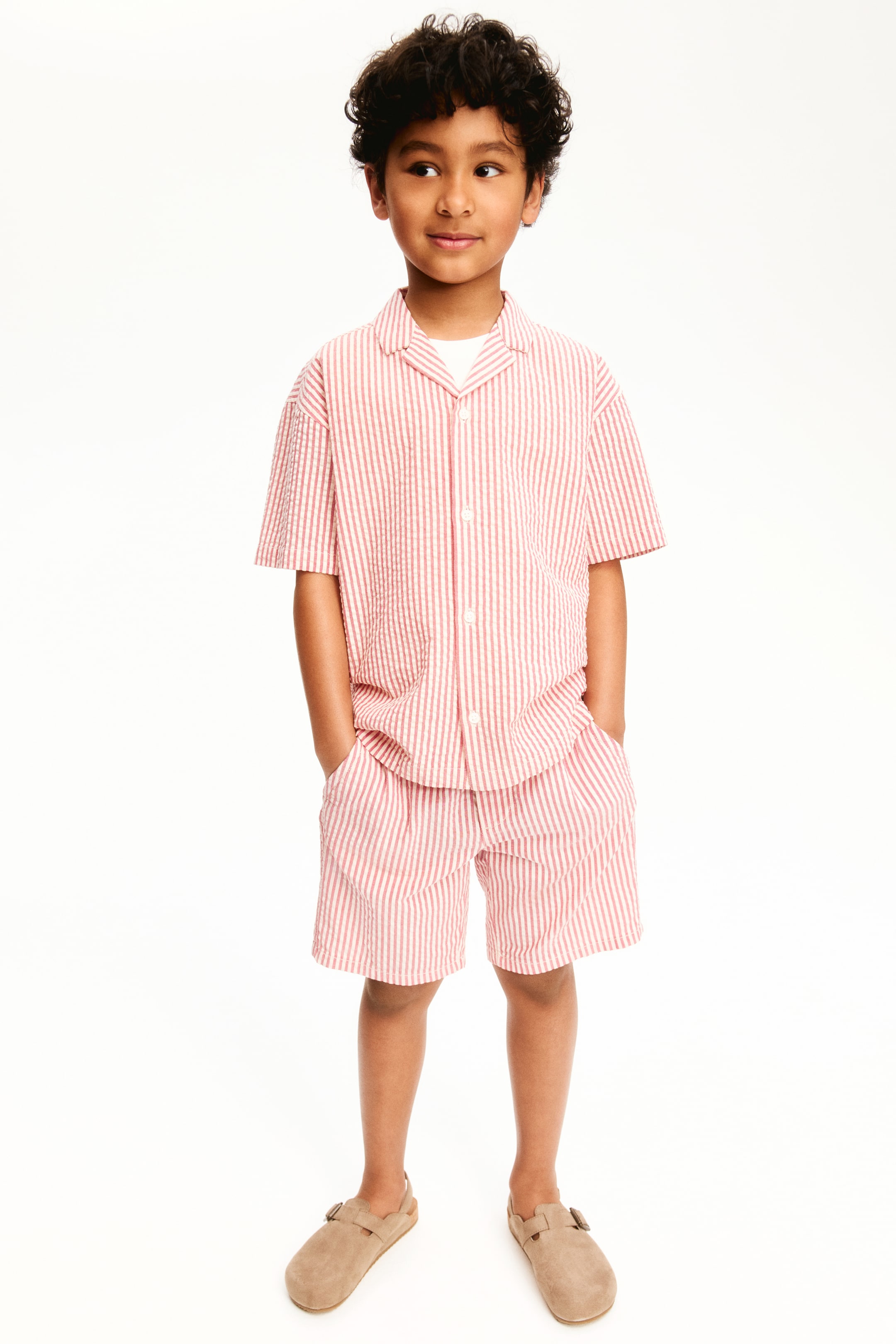 Größeres Bild ansehen: Ein rot-weiß gestreiftes Seersucker-Kurzarmhemd mit einem Camp-Kragen und weißen Knöpfen wird über einem weißen Unterhemd getragen und mit passenden gestreiften Shorts mit Taschen kombiniert. Auch tanfarbene Schnallen-Clogs sind sichtbar.