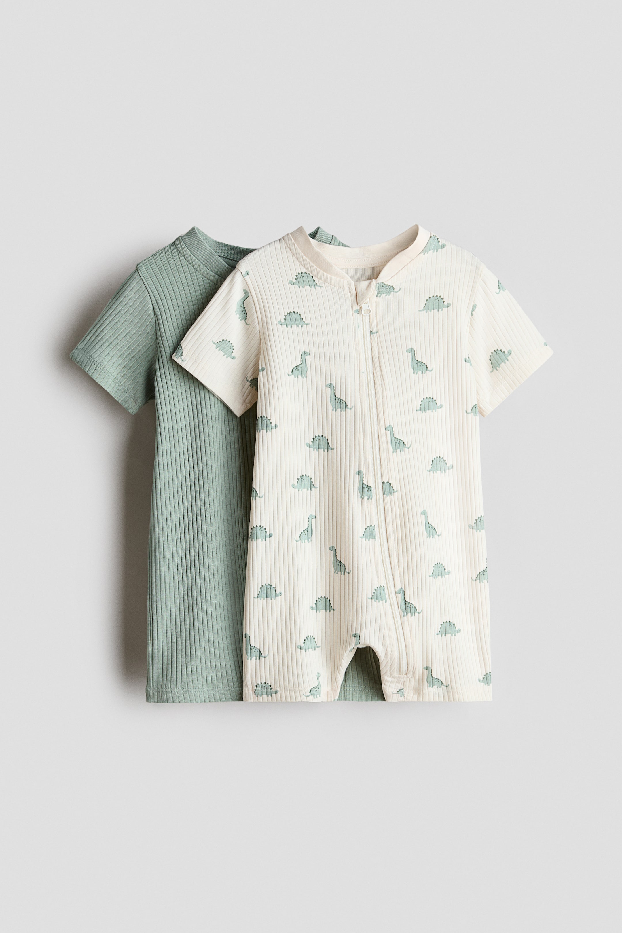 Xem ảnh lớn hơn: Hai bộ đồ ngủ ngắn tay được trưng bày từ phía trước, làm từ chất liệu cotton jersey gân mềm mại. Bộ đồ ngủ phía trước có màu trắng ngà với họa tiết khủng long màu xanh nhạt, có khóa kéo chạy dọc phía trước và một bên chân, với miếng bảo vệ cằm chống trầy xước che phủ phía trên. Một bộ đồ ngủ màu xanh nhạt trơn được xếp lớp phía sau; cả hai bộ đồ ngủ đều có chân ngắn và tay ngắn.