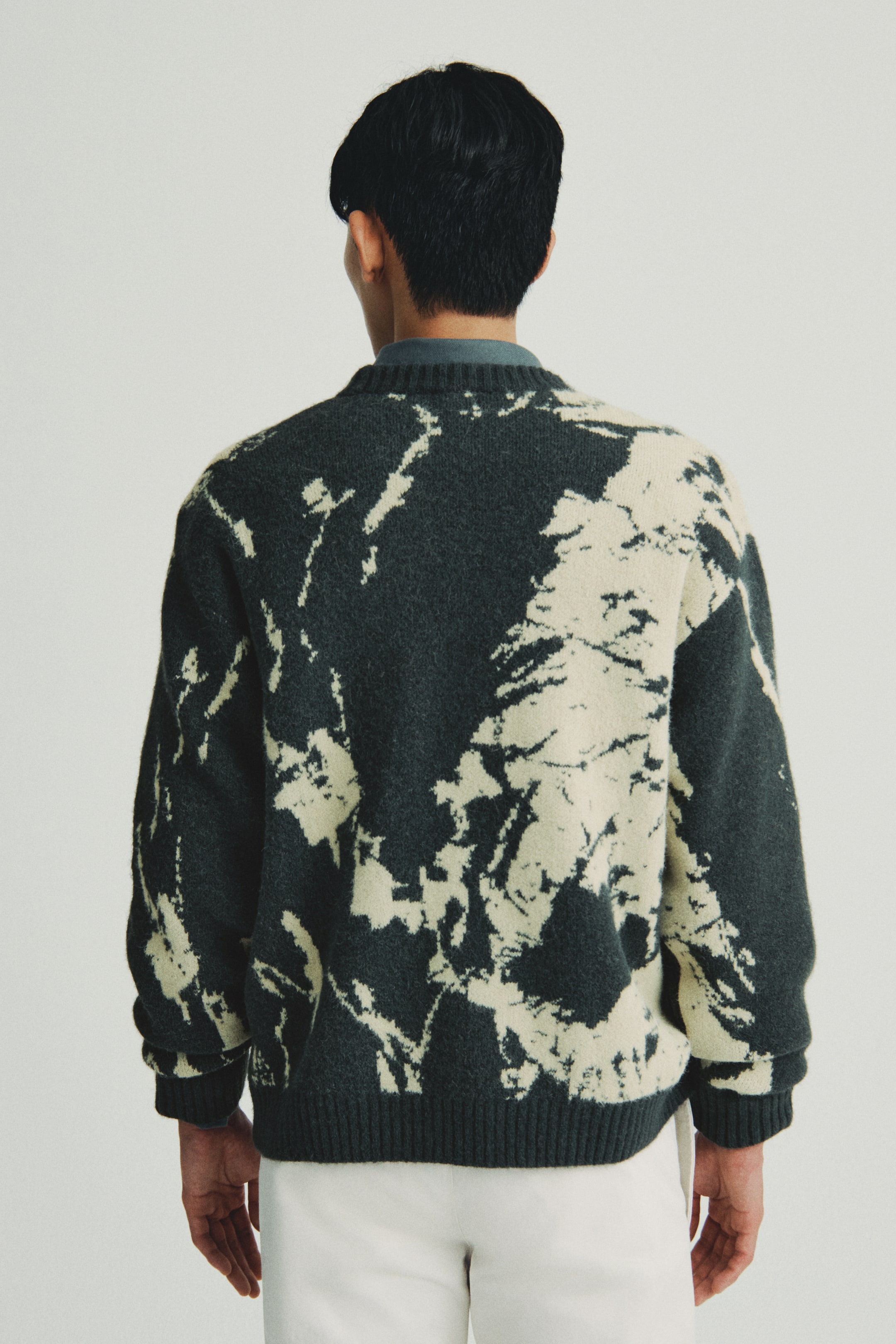 이미지 확대 보기: 짙은 회색 니트 스웨터가 뒷모습을 보여주고 있으며, 베이지색 추상 패턴이 특징입니다. 라운드 넥 디자인, 골지 소매 및 골지 밑단으로 디자인되어 있으며, 청회색 칼라 셔츠 위에 레이어링되어 있습니다.