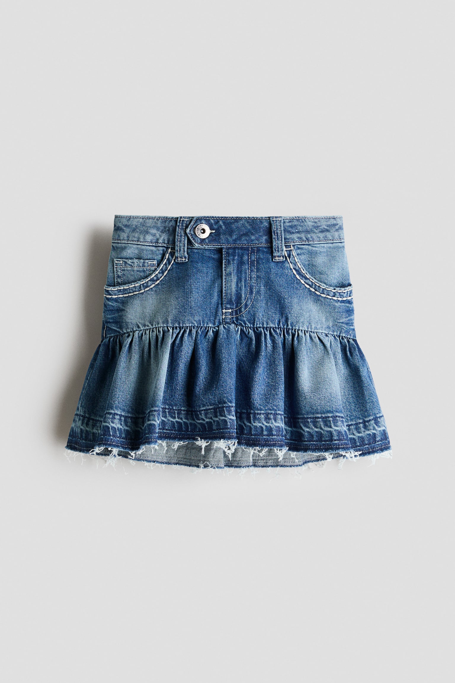 A blue denim skirt is positioned front-facing, exhibiting a medium wash with lighter fading. It features a button fastening, zip fly, belt loops, and two front pockets with white contrast topstitching. A gathered ruffle tier forms the lower section, ending in a distressed, frayed hem. - 1