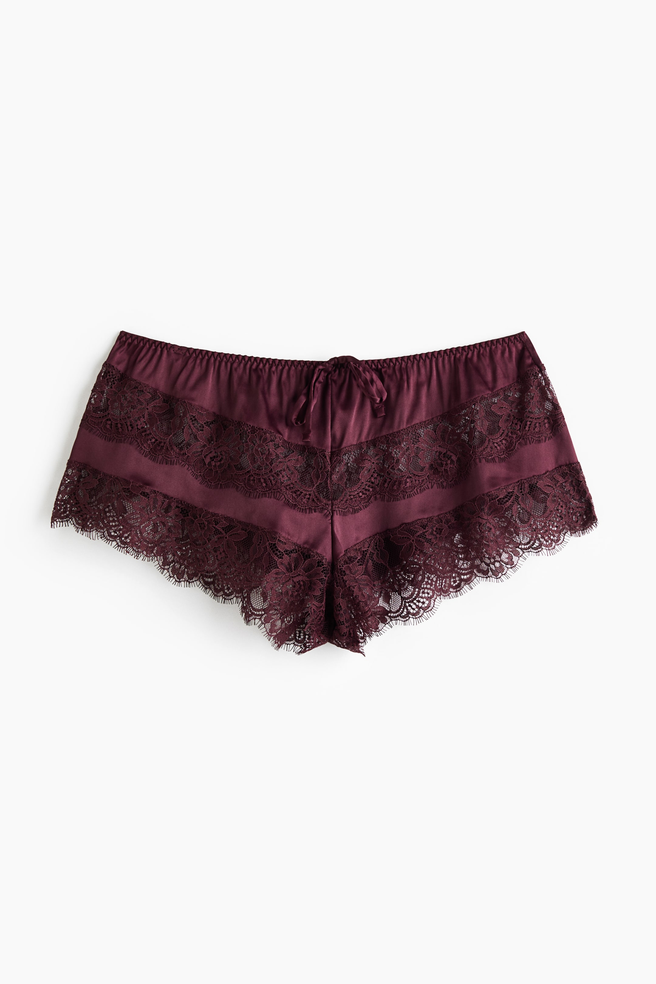 Agrandir l'image: Une paire de shorts tap en satin bordeaux est présentée de face, mettant en valeur une taille élastiquée avec un petit nœud centré. Le modèle présente des panneaux alternés de dentelle florale transparente assortie et une bordure en dentelle festonnée le long des ouvertures de jambes.