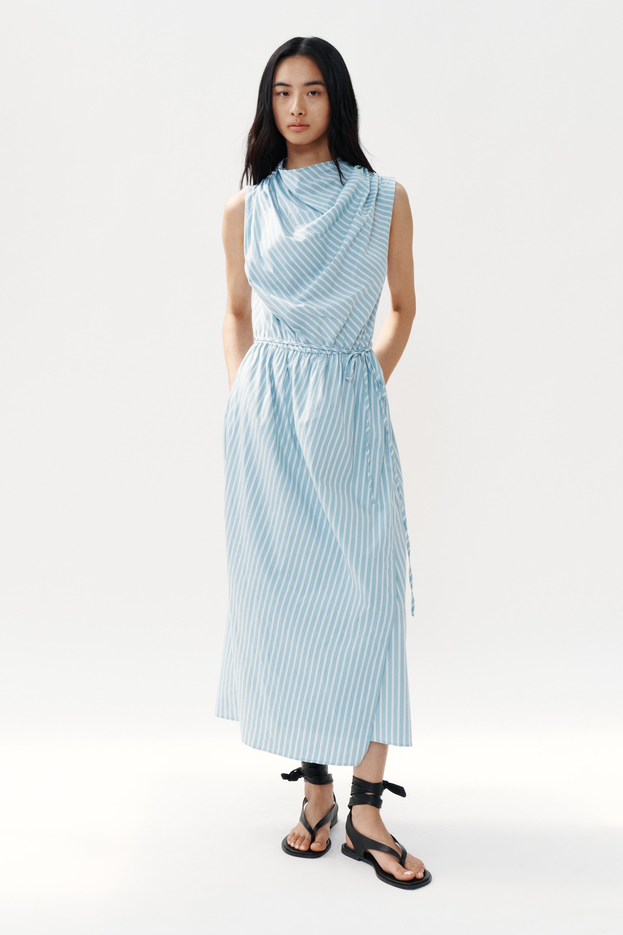 View larger image: A light blue and white striped sleeveless maxi dress features a draped cowl neckline and a cinched waist with a tie-belt. The dress flows to the mid-calf and includes visible side pockets, styled with black flat thong sandals that have ankle-wrap ties.
