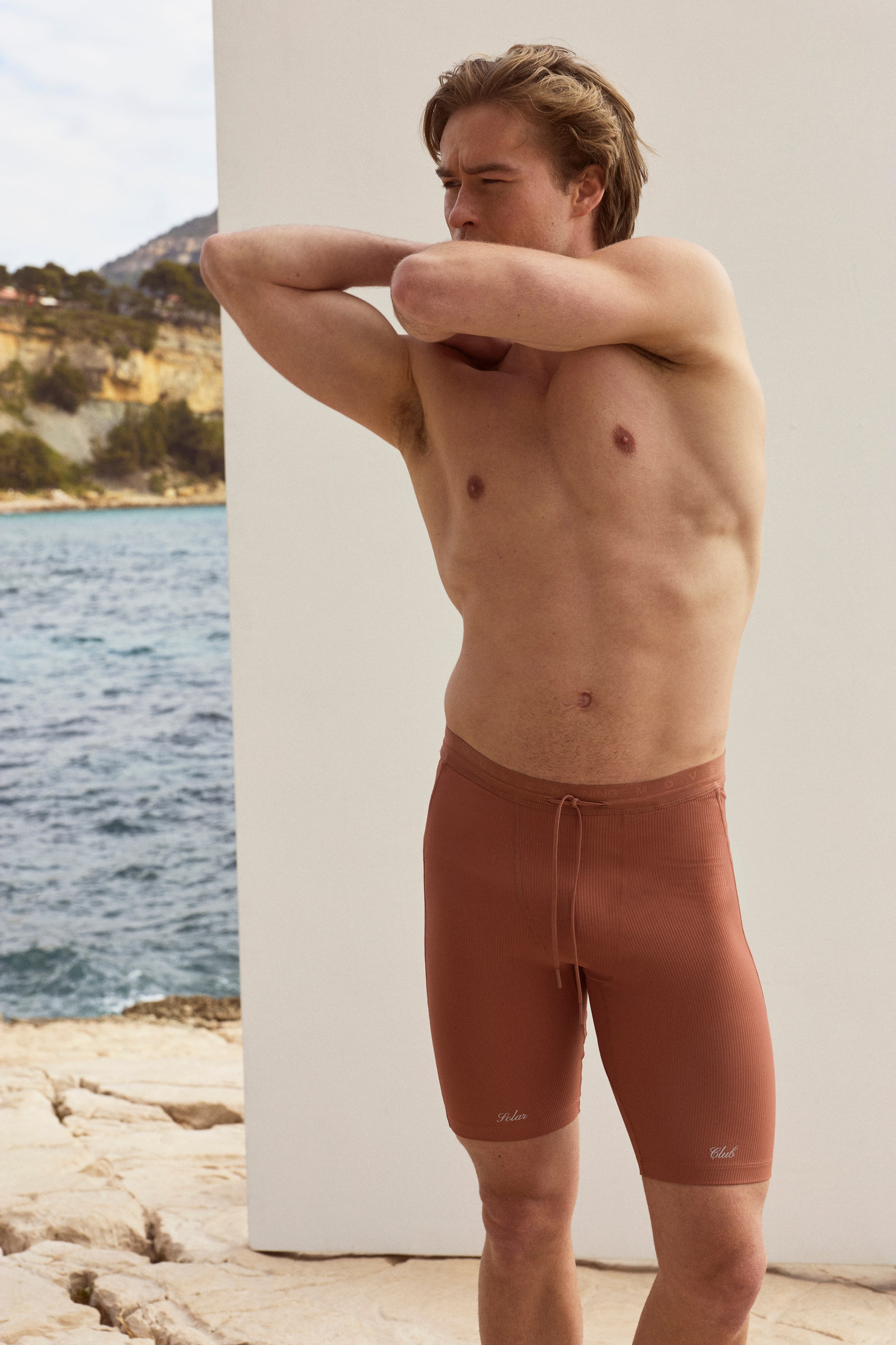 Agrandir l'image: Un short en maille côtelée couleur terracotta s'ajuste parfaitement sur le haut des cuisses, doté d'une taille élastiquée avec un cordon de serrage visible à l'avant au centre. De petits textes brodés, "Solar" et "Club," apparaissent sur les cuisses gauche et droite.