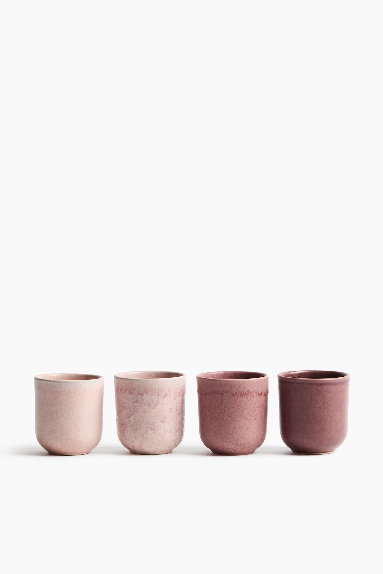 Quattro tazze in gres senza manico sono disposte una accanto allâaltra, rivolte frontalmente, e mostrano una sfumatura di colore dal rosa pallido al prugna intenso. Ogni tazza presenta una forma rustica e arrotondata con una smaltatura reattiva variegata unica, che crea un aspetto puntinato e materico.