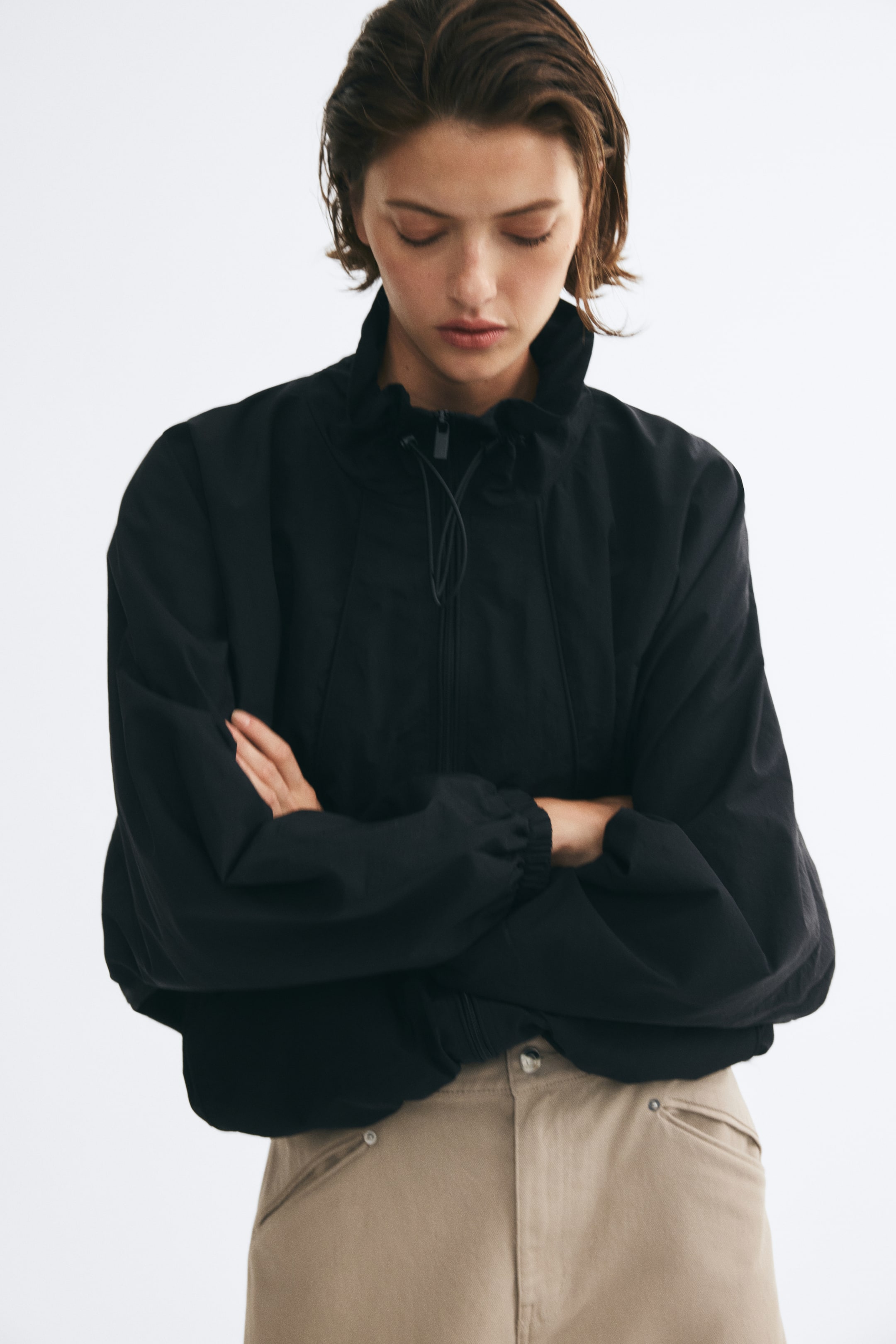 View larger image: The model is wearing a black oversized nylon jacket and standing with arms crossed. The jacket features a high funnel collar with a drawstring, balloon sleeves with elasticated cuffs, and a front zipper, and is paired with high-waisted beige trousers.