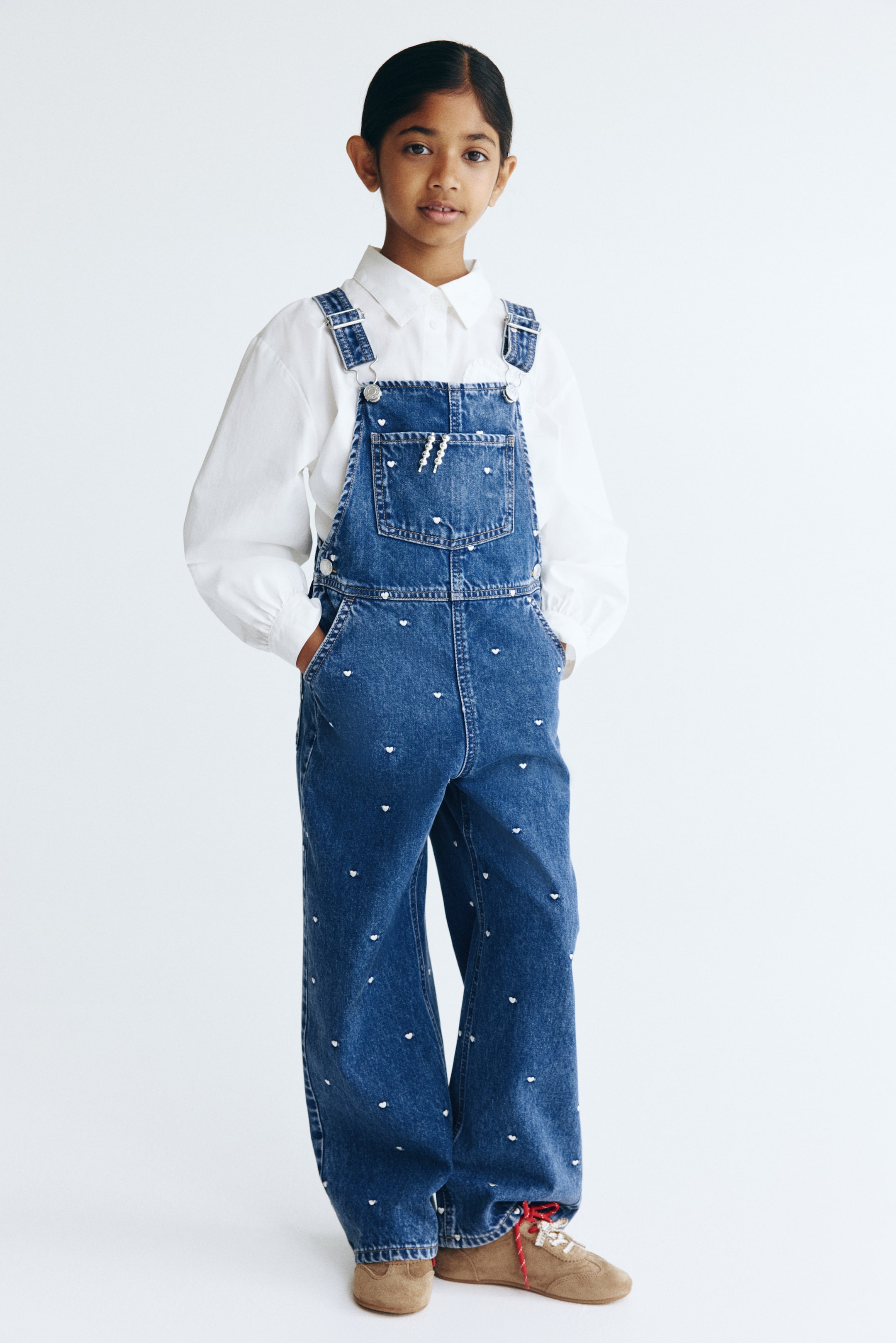 Grotere afbeelding bekijken: Voorzijde tuinbroeken in blauw patroon katoen denim met kleine witte hartjes overal, verstelbare schouderbanden met metalen sluitingen, een borstzakje met een witte bliksemschicht en kralen, zij knopen, diagonale zijzakken en rechte pijpen.