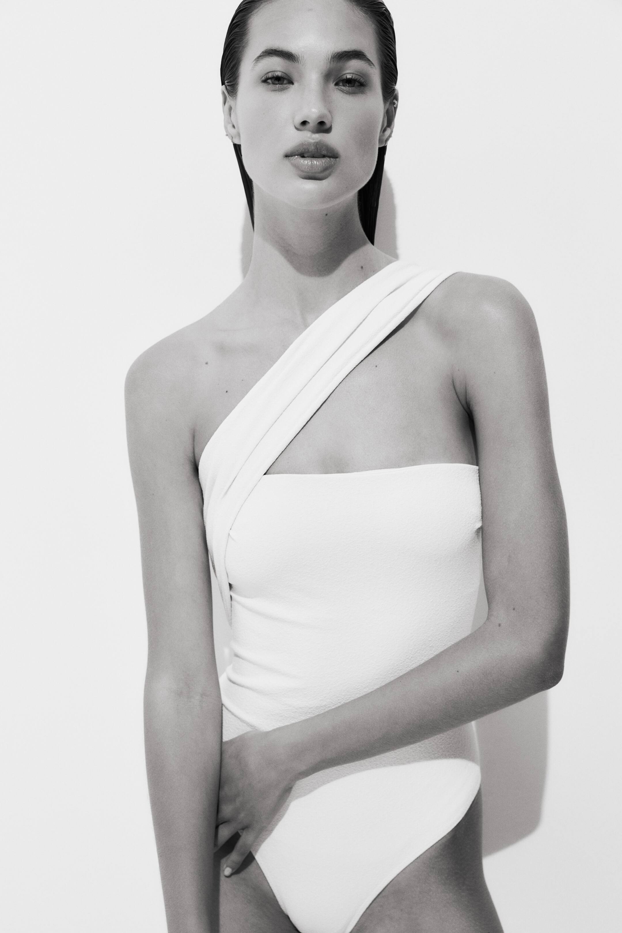 View larger image: A light-colored one-piece swimsuit featuring an asymmetrical design, with a wide strap crossing from the left shoulder to a bandeau-style bodice, made from subtly textured fabric.