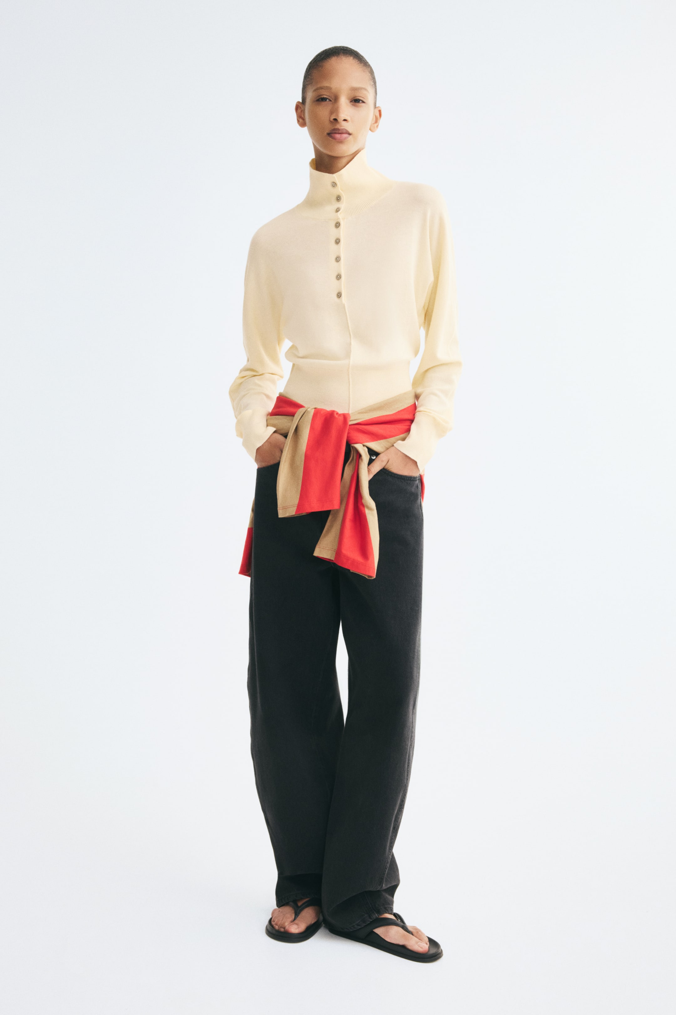 View larger image: A cream-coloured, fine-knit sweater is shown from the front, featuring a high neck, a button placket with pearly buttons, and long sleeves with ribbed cuffs. The slim-fit sweater is worn tucked into dark trousers, with another knit garment tied around the waist.