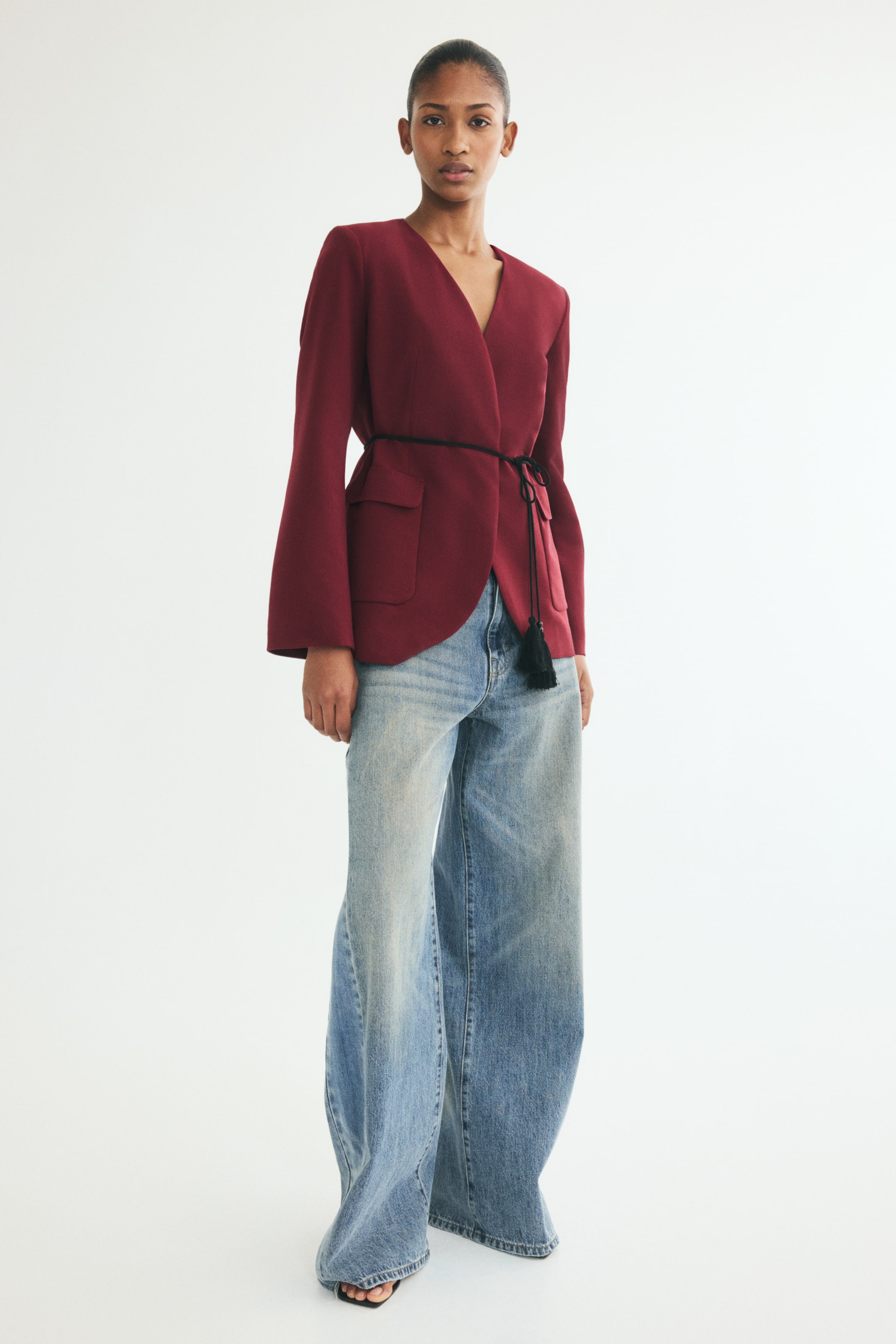 View larger image: This burgundy, lapelless blazer is presented from a three-quarter front angle, featuring structured shoulder pads, a deep V-neckline, and front flap pockets. The blazer is styled closed with a thin black twisted rope tie belt with tasseled ends at the waist, worn over wide-leg light blue jeans.