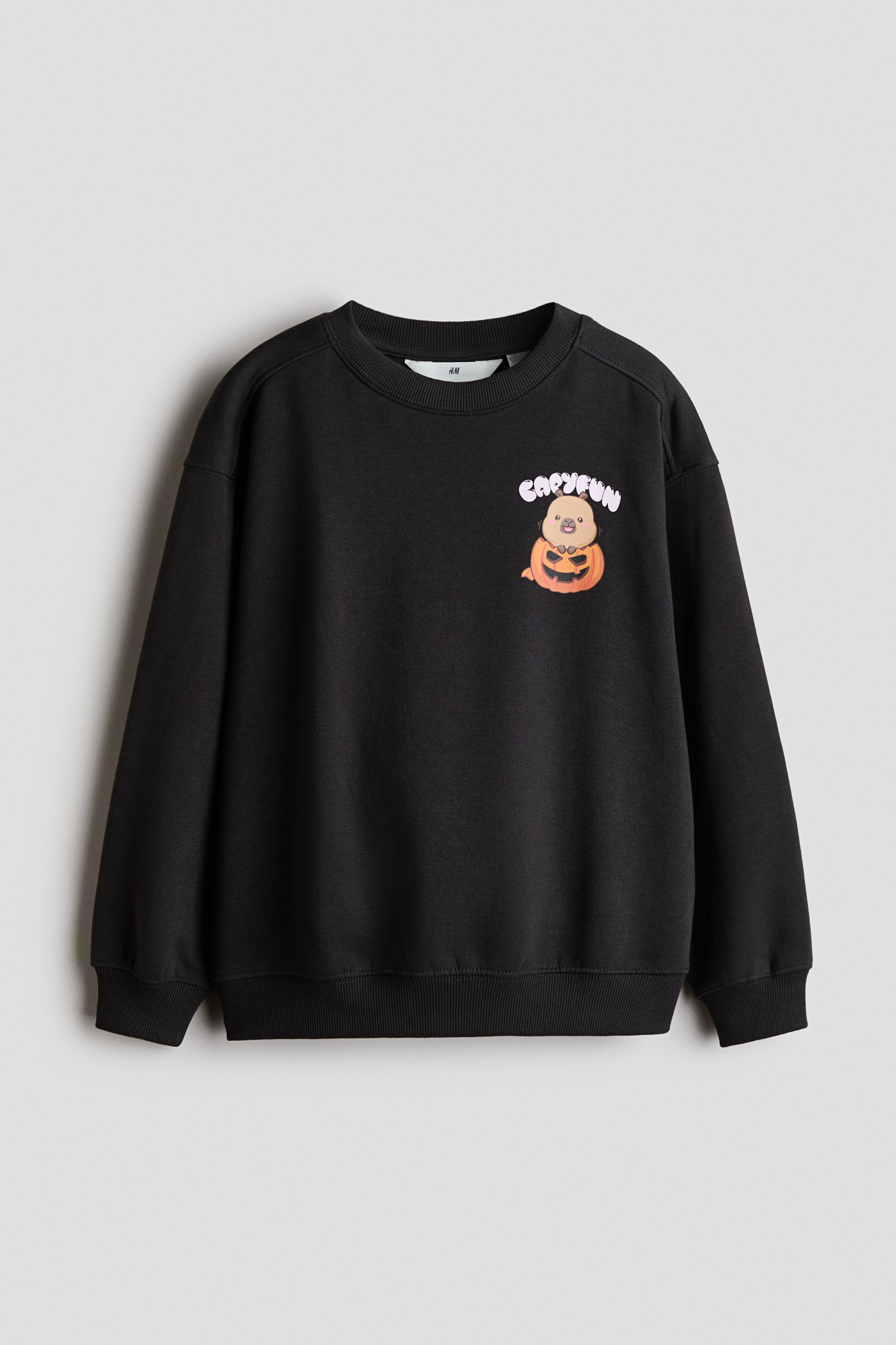 Agrandir l'image: Un sweat-shirt noir à coupe ample est présenté de face, avec un col rond côtelé, des épaules tombantes et des bord-côtes aux poignets et à l’ourlet. Un motif imprimé est visible sur le devant, représentant une créature brun clair assise dans une citrouille d’Halloween orange, avec le texte « CAPYFUN » au-dessus.