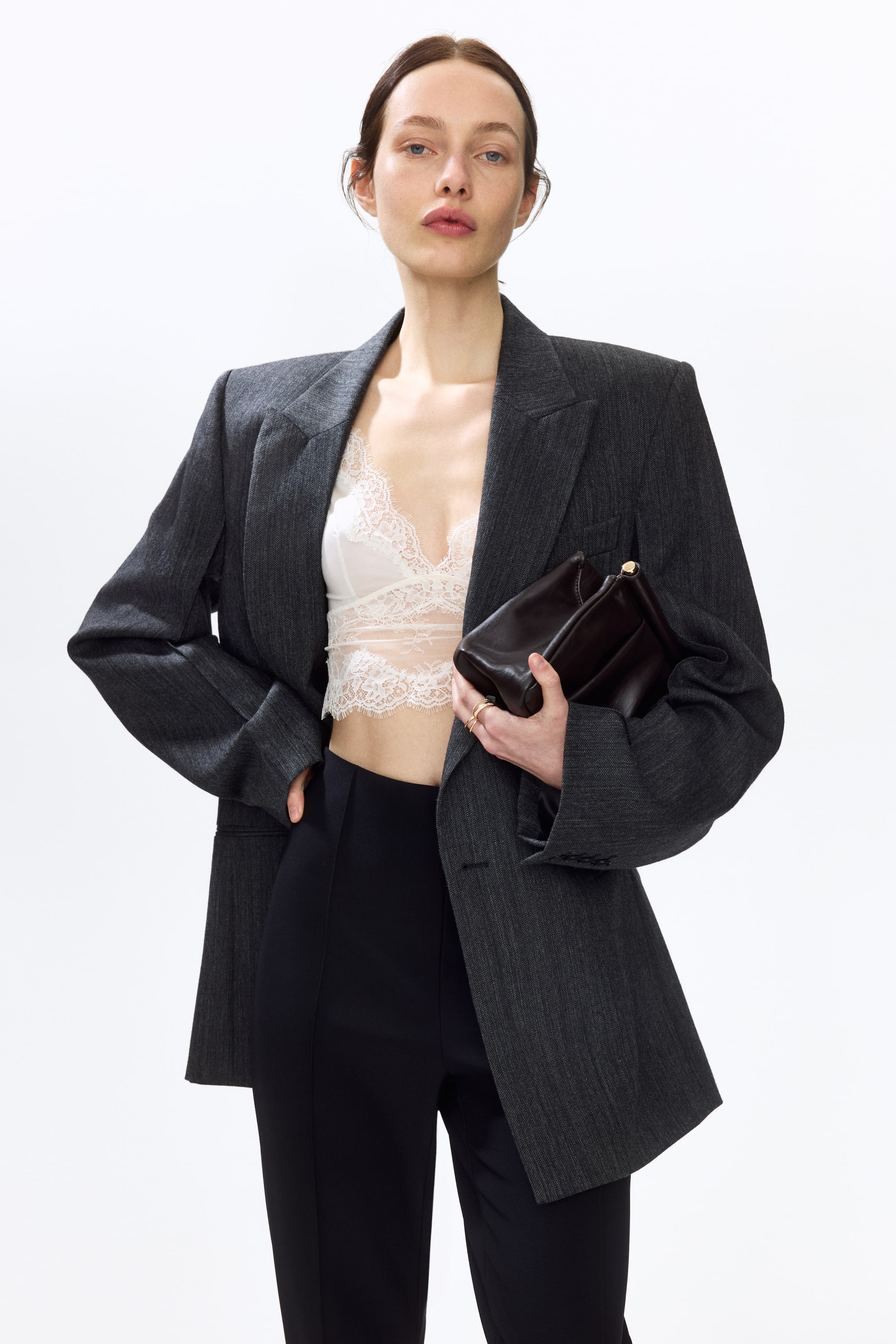 View larger image: A dark grey pinstripe blazer is showcased from a front-facing angle, featuring broad, structured shoulders, notch lapels, and a single-button fastening. It is styled open in a relaxed fit over a white lace bralette.
