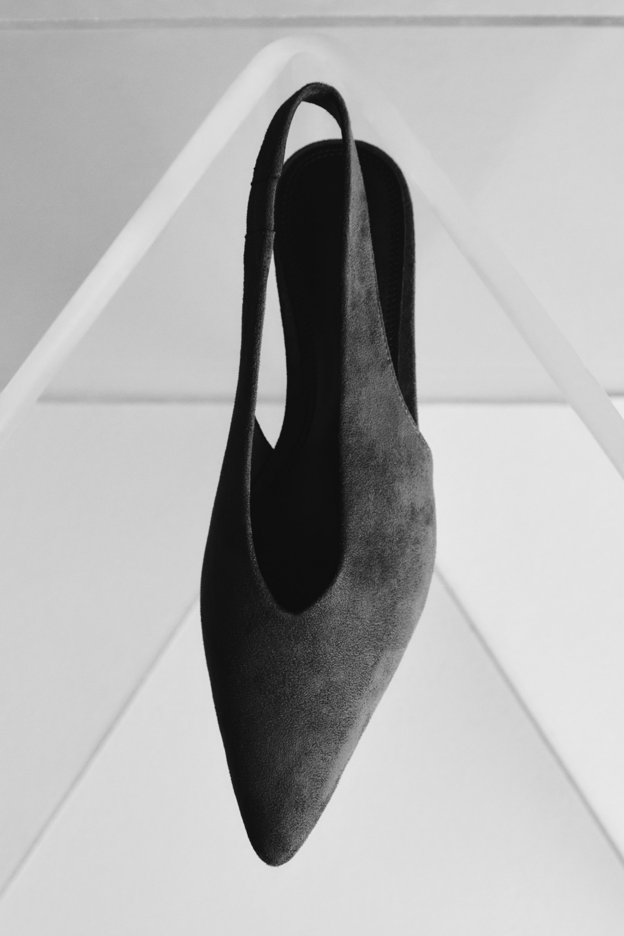 Grotere afbeelding bekijken: Een donkergrijze slingback schoen staat iets naar rechts gericht, waarbij de spitse neus en de diepe V-vormige vamp worden getoond. Een zichtbare slingback band buigt over de bovenkant, en de schoen is gemaakt van een getextureerd, suède-achtig materiaal.