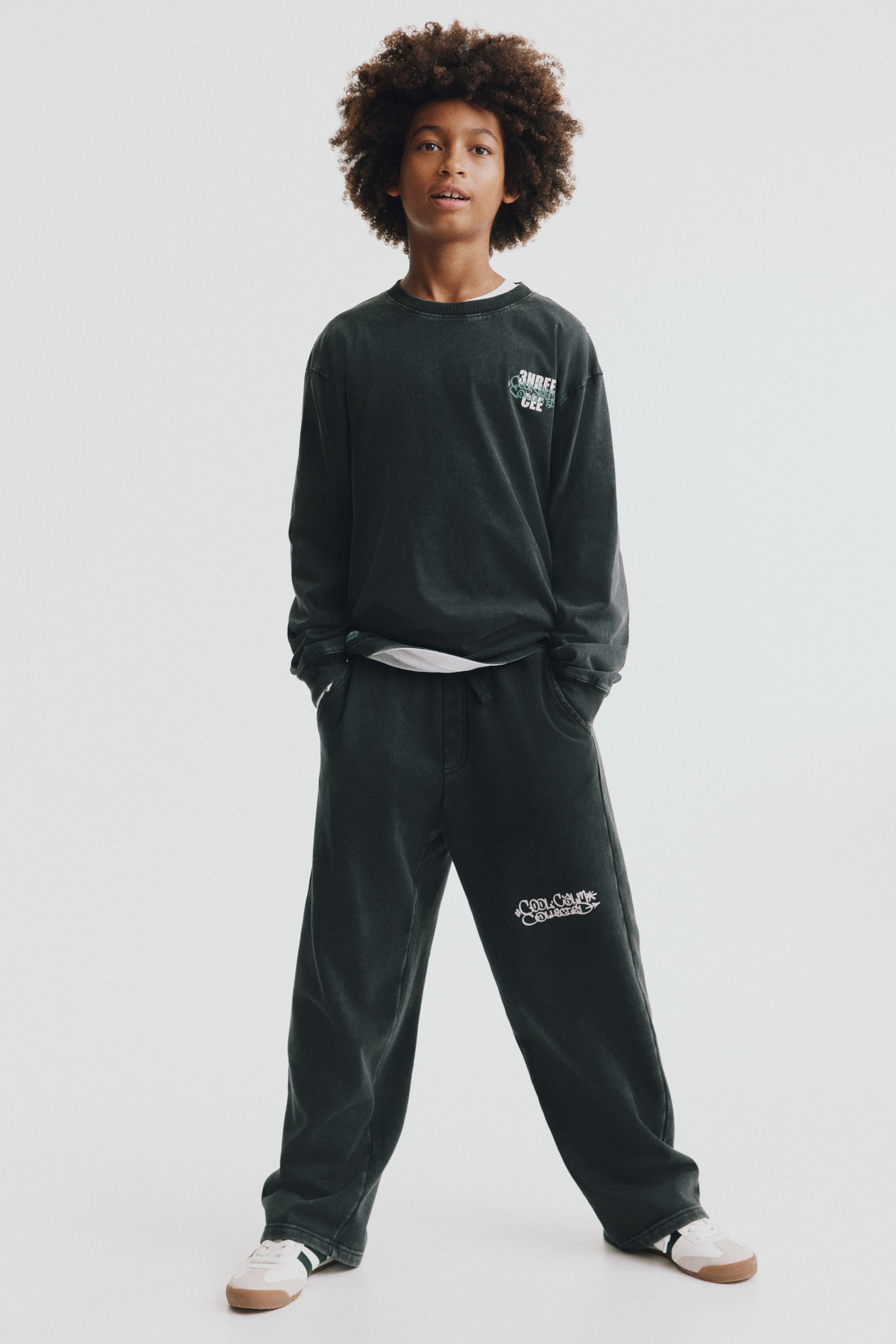 View larger image: A child is wearing a front-facing, dark green matching set consisting of a long-sleeve jersey top and straight-leg track pants. The top features a ribbed crew neckline, ribbed cuffs, and a small white printed design on the chest, styled over a layered white T-shirt. The matching track pants feature side pockets and a white graphic print on the left thigh.