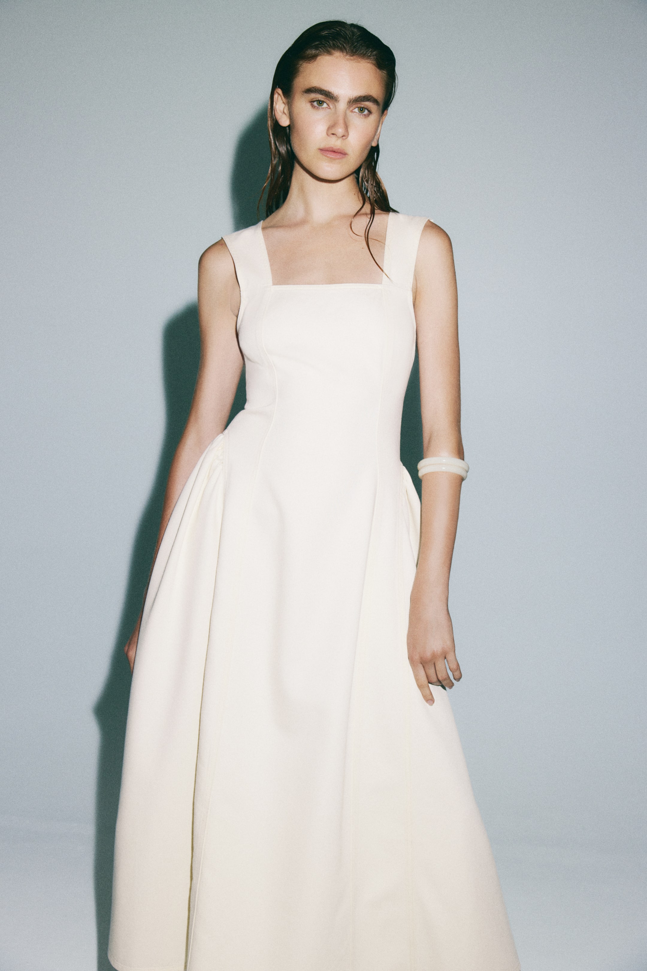 View larger image: A calf-length cream dress, with a square neckline, wide shoulder straps, a fitted bodice, and a flared skirt showing visible seams down the front, is being worn by the model. Two light-coloured bangles are also being worn on the model’s right wrist.