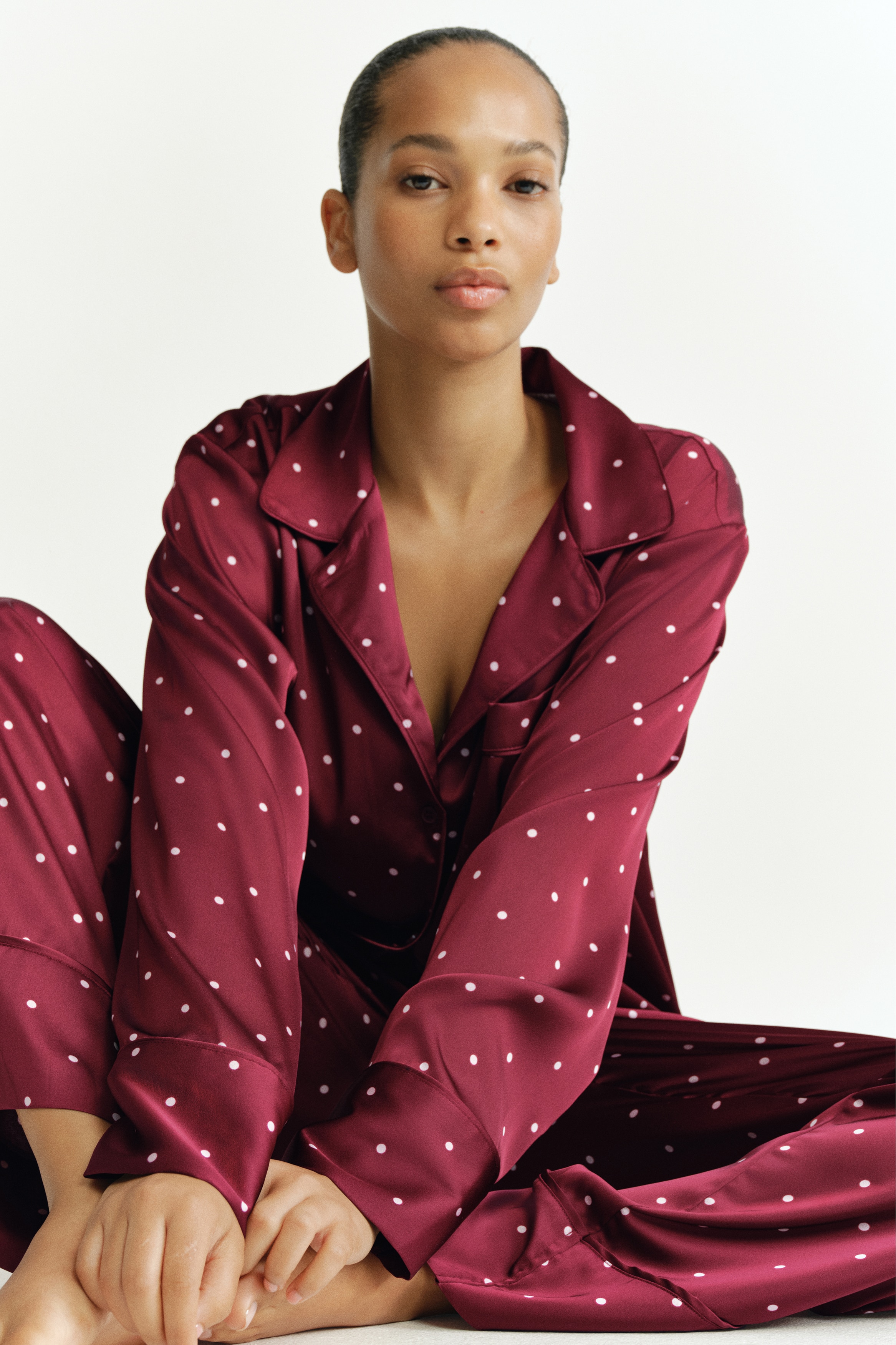 Un completo pigiama in raso color bordeaux con fantasia a pois bianchi Ã¨ appeso frontalmente. La camicia a maniche lunghe presenta un colletto resort, apertura frontale con bottoni e una tasca singola sul petto, mentre i pantaloni coordinati a gamba larga pendono dritti sotto.