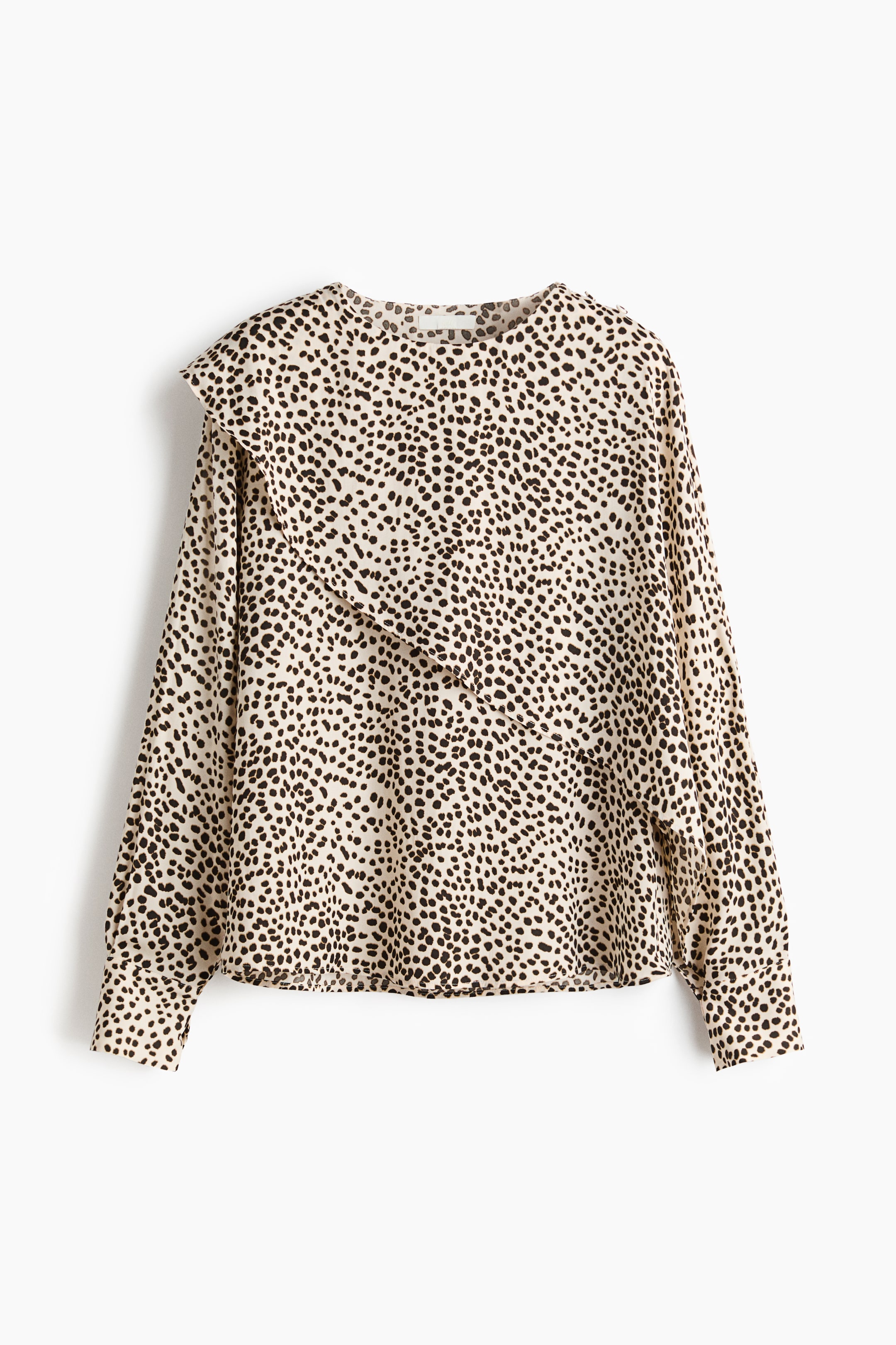 Se større billede: En frontvendt bluse viser et beige stof med et sort plettet mønster. Den har en rund halsudskæring, lange ærmer med knappede manchetter og en stor flæsedetalje, der strækker sig fra venstre skulder tværs over fronten.