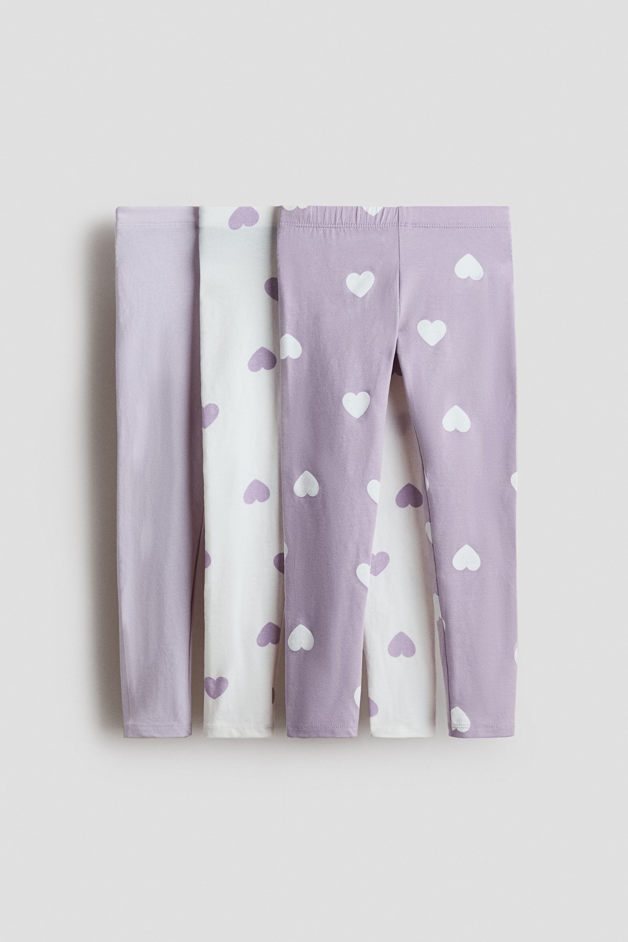 View larger image: Four pairs of leggings crafted from soft cotton jersey with an elastic waistband are displayed, showcasing their front view. They come in a variety of designs: a solid light purple, two white pairs adorned with purple hearts, and one light purple pair featuring white hearts.