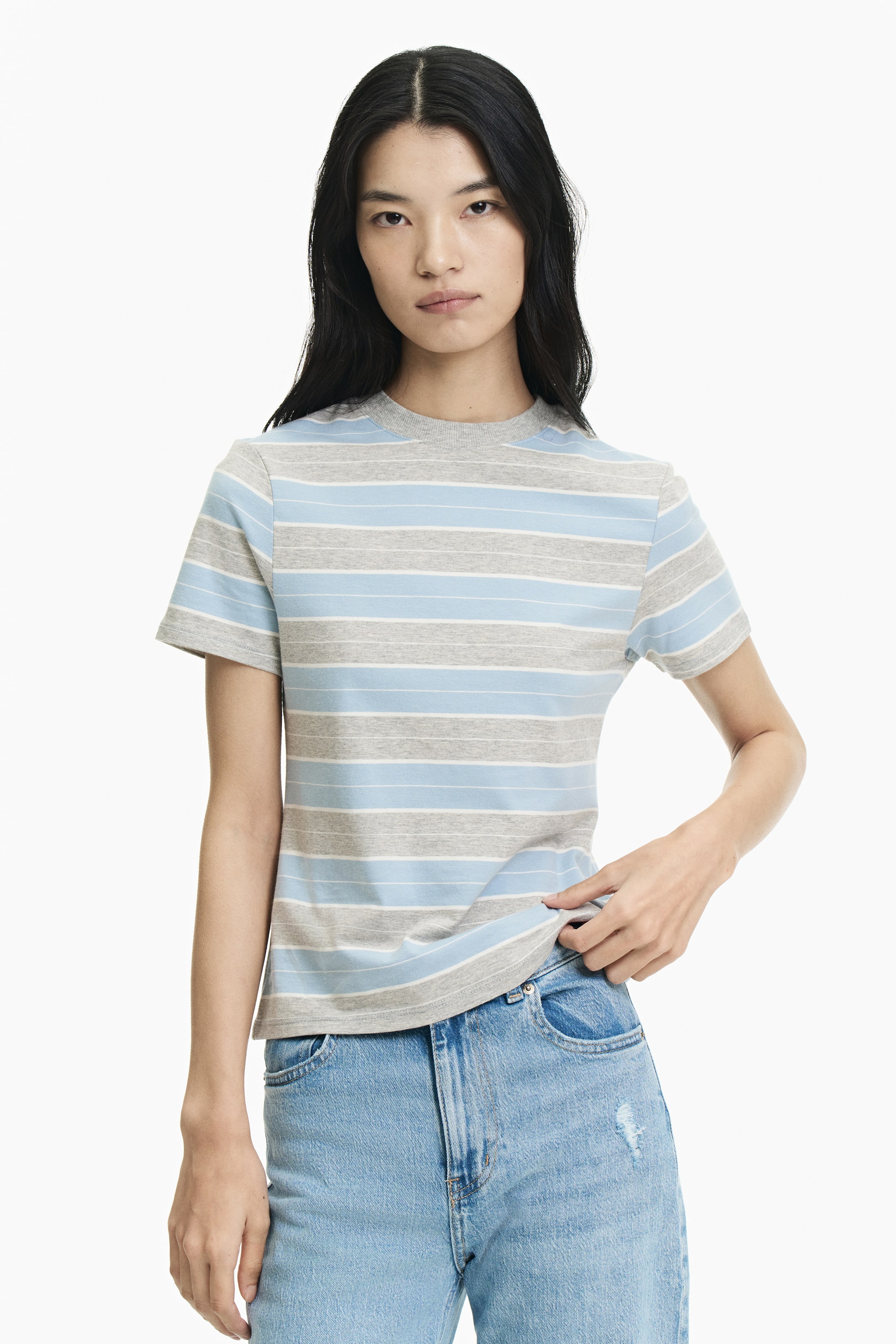 拡大画像を表示: モデルは、グレー、ライトブルー、細いホワイトの横ストライプが入った半袖Tシャツを着用しており、リブ仕上げのラウンドネックが特徴です。Tシャツのストレートカットの裾を持ちながら、ライトブルーのジーンズも着用しています。