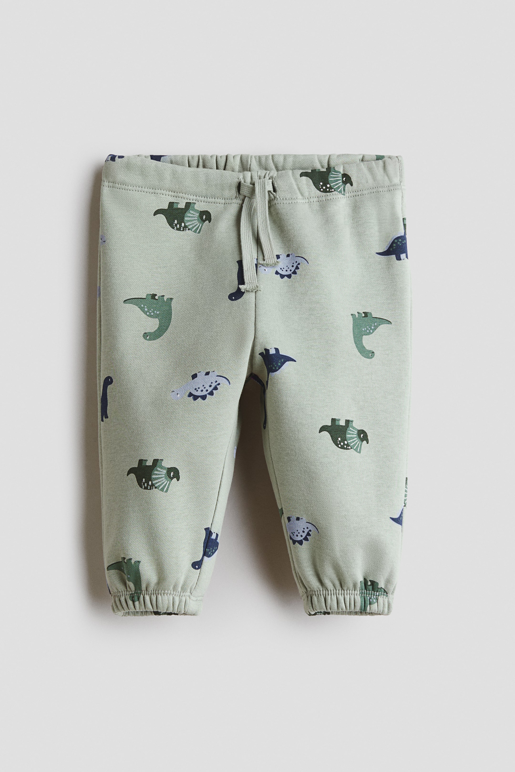 Büyük resmi görüntüle: Jogger pantolonlar önden sergilenmekte olup, tüm yüzeyinde dinozor baskısı bulunan açık yeşil sweatshirt kumaştan üretilmiştir. Ayarlanabilir, lastikli bir bel kısmı, dekoratif bağcık detayı ve paçalarda kapalı elastik yapısı öne çıkarılmaktadır.