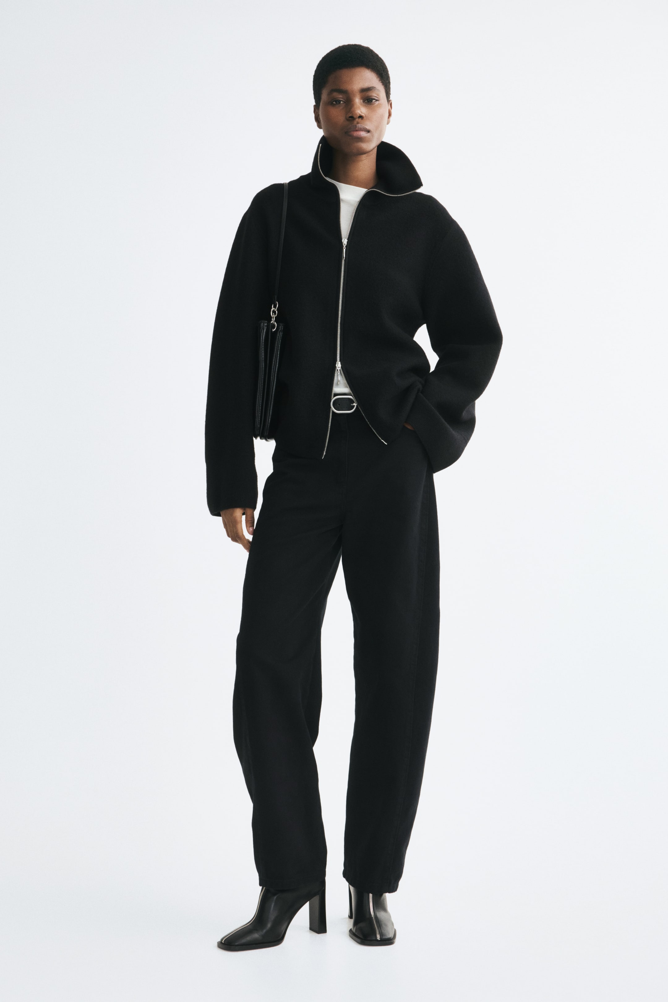 View larger image: A black fine-knit wool cardigan with a high collar, dropped shoulders, and a silver-tone two-way front zipper is worn by the model. The cardigan is styled partially unzipped at the collar and hem, layered over a white top, and paired with black wide-leg trousers and a black shoulder bag.