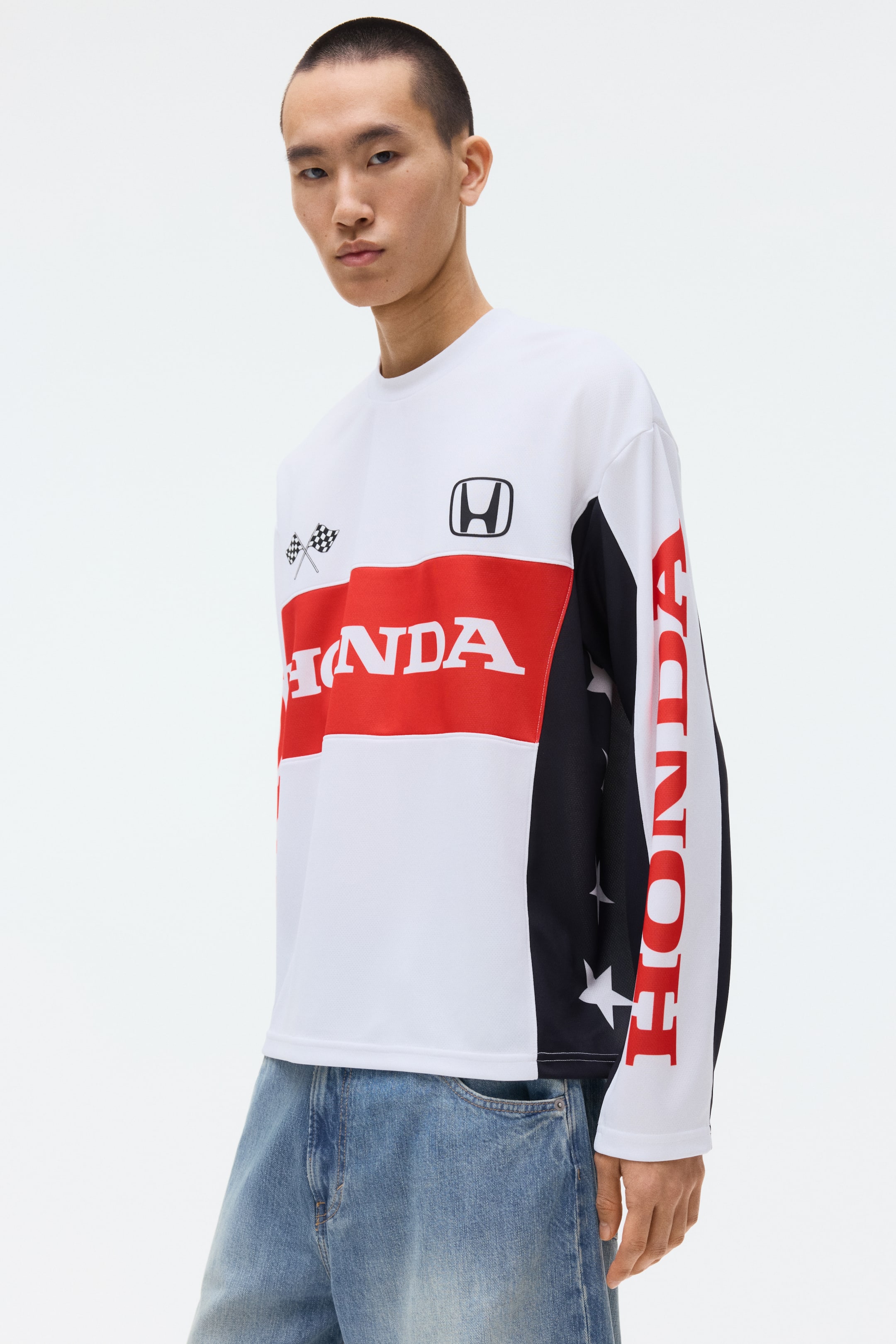 拡大画像を表示: 長袖の白いトップスは、胸に赤い水平ストライプと白い「HONDA」テキストが特徴で、着用者の右上胸部には黒いホンダのロゴ、左上胸部には交差したチェッカーフラッグが描かれています。着用者の左側のパネルと袖には、白い星があしらわれた黒いデザインが施されており、右袖には縦に赤い「HONDA」テキストが表示されています。