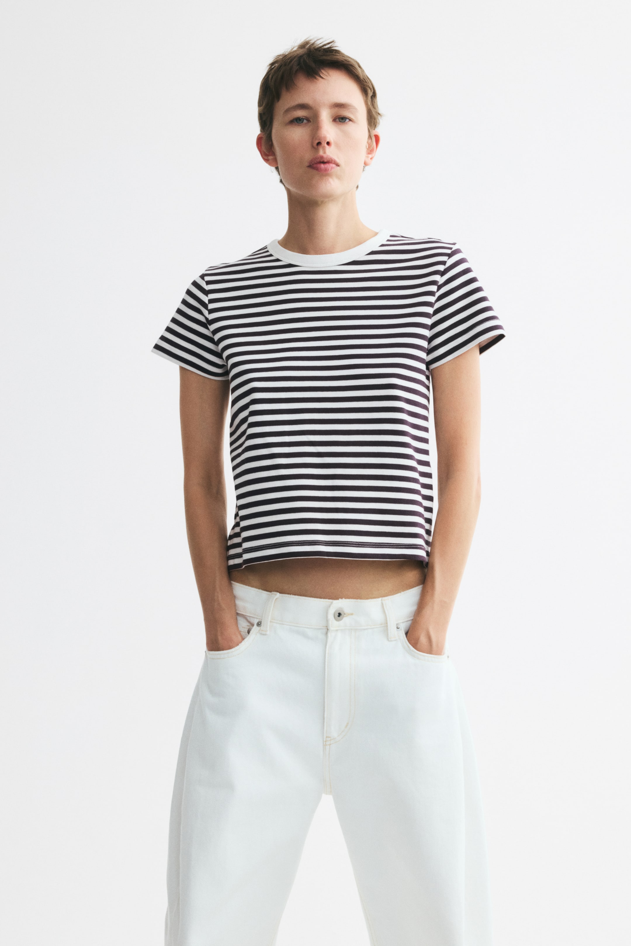 View larger image: A fitted T-shirt with black and white horizontal stripes, a ribbed neckline, and a straight, cropped hem is worn by the model. The model is standing with hands in the light-coloured trouser pockets, showcasing the T-shirt's fit and length.