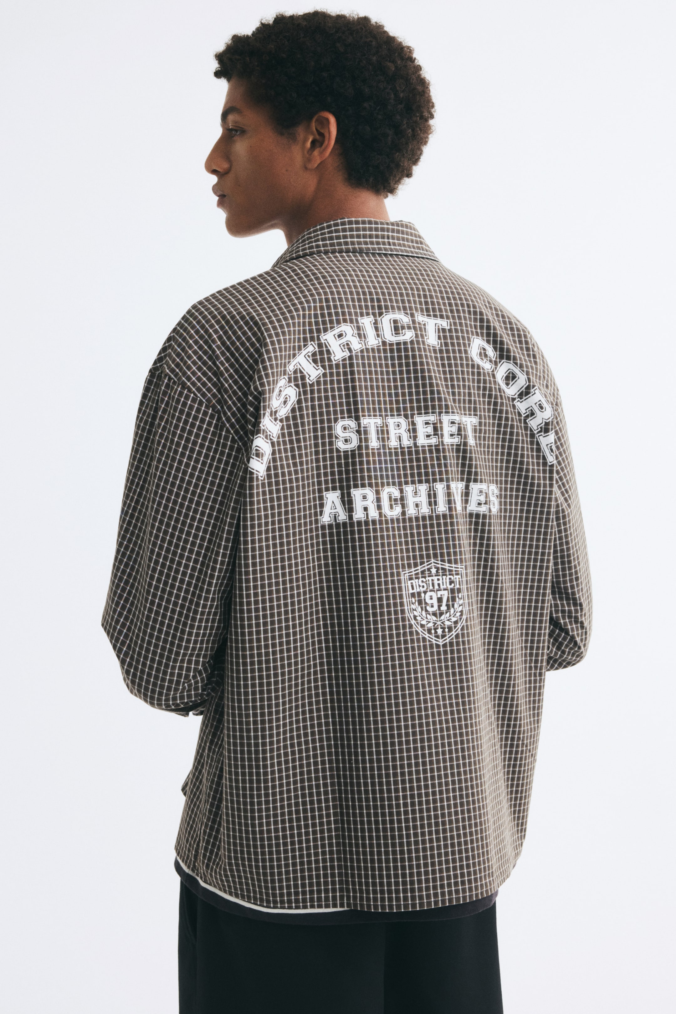 View larger image: A long-sleeved, relaxed-fit shirt in a brown and white grid pattern is worn by the model, who is turned away from the camera. The shirt features dropped shoulders, a straight hem, and bold white text reading "DISTRICT CORE STREET ARCHIVES" printed on the back above a shield graphic, and is styled with dark trousers.