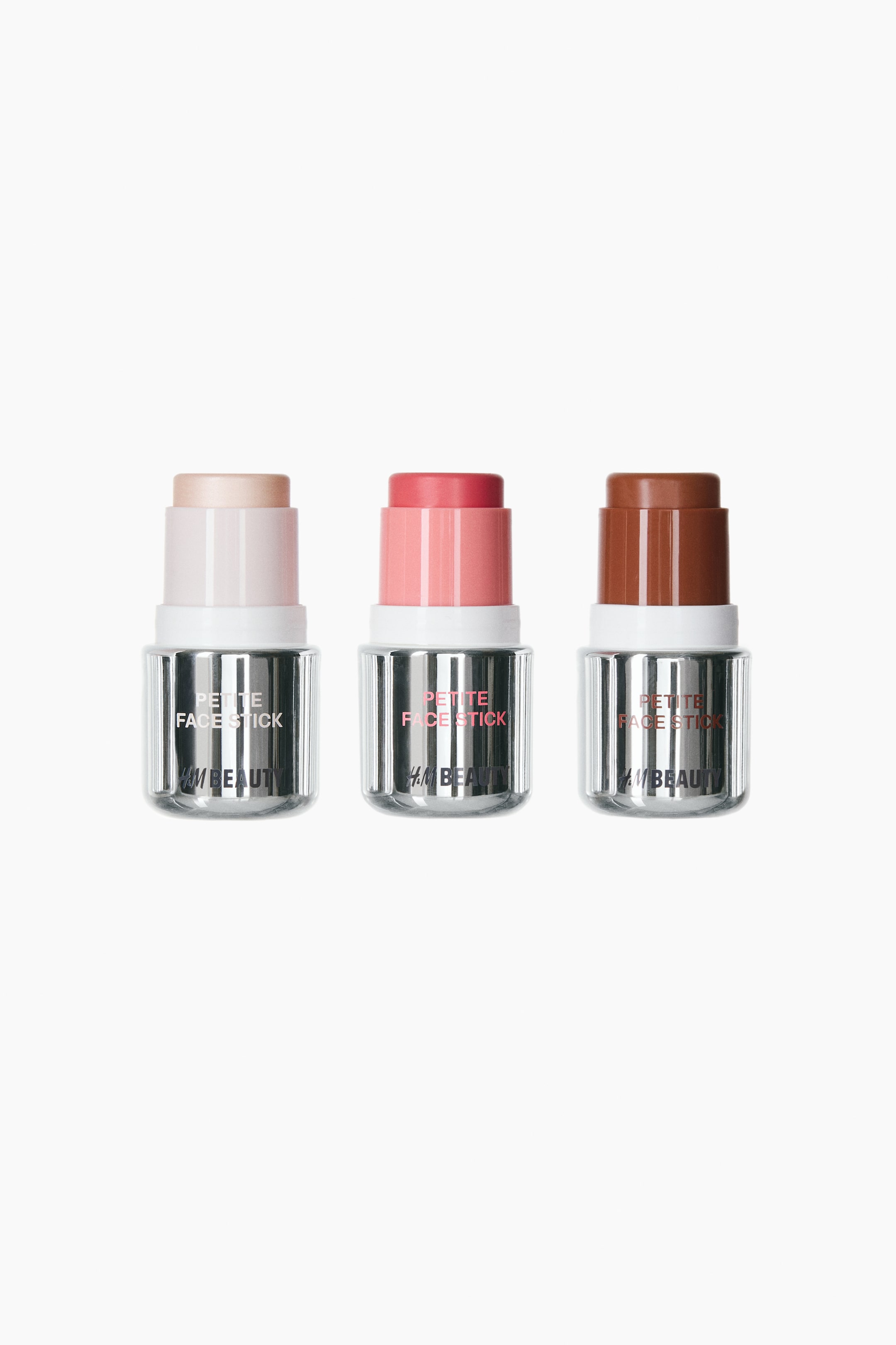 View larger image: Three face sticks stand front-facing in a row, each displaying its product extended from the top. The left stick shows a light beige highlighter, the middle stick a coral pink blusher, and the right stick a medium brown bronzer. Each stick features a silver-toned ribbed base with "PETITE FACE STICK" and "H&M BEAUTY" printed on the front.