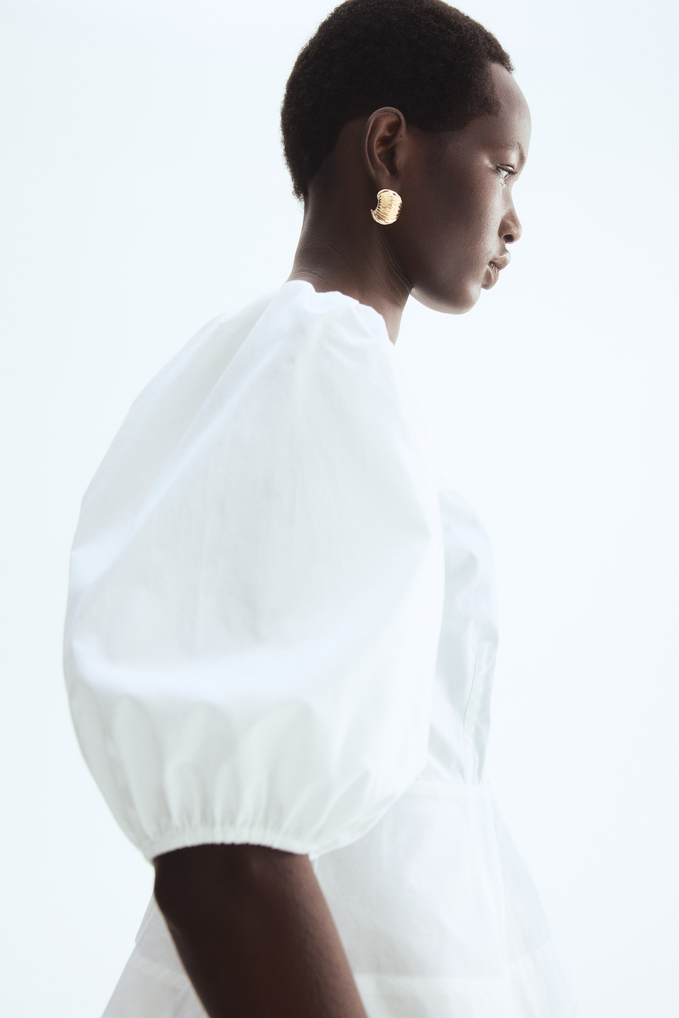 View larger image: The model is wearing a white, short, fitted blouse crafted from crisp cotton poplin, featuring elbow-length puff sleeves and a flared peplum.