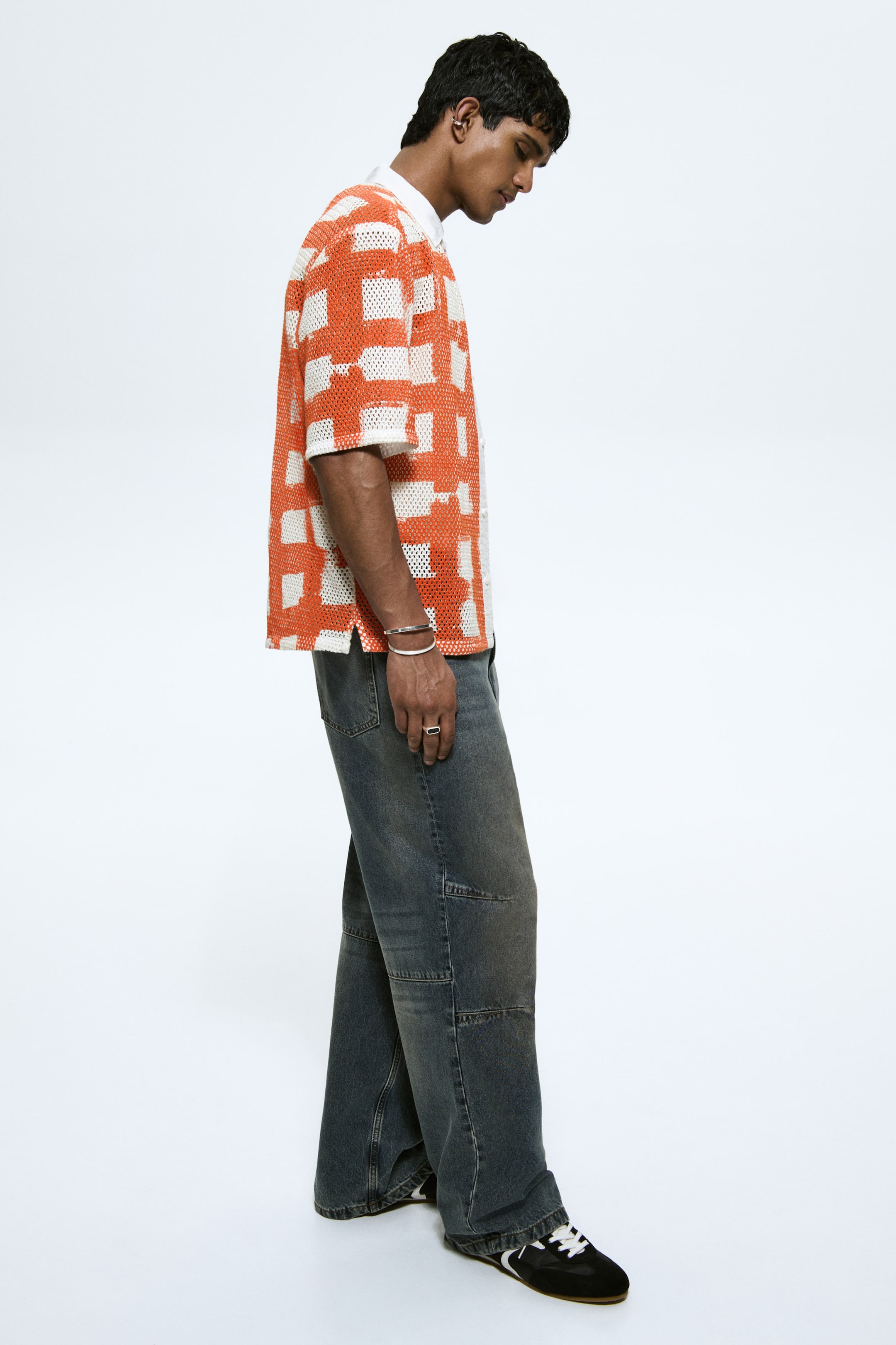 View larger image: A short-sleeved, collared shirt in an orange and white chequered pattern, crafted from perforated mesh fabric, shown in a relaxed fit with side slits from a side angle, layered over a white collared shirt.