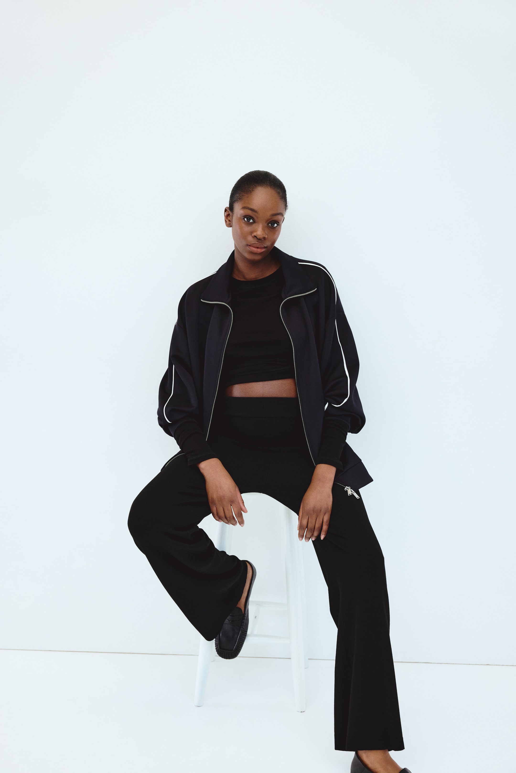 View larger image: A black tracksuit features a tracksuit jacket with a full-zip front, a stand-up collar and white piping along the shoulders and outer sleeves. The jacket is worn open over a black crop top and paired with matching wide-leg tracksuit bottoms, which display a zip detail on the lower left leg.