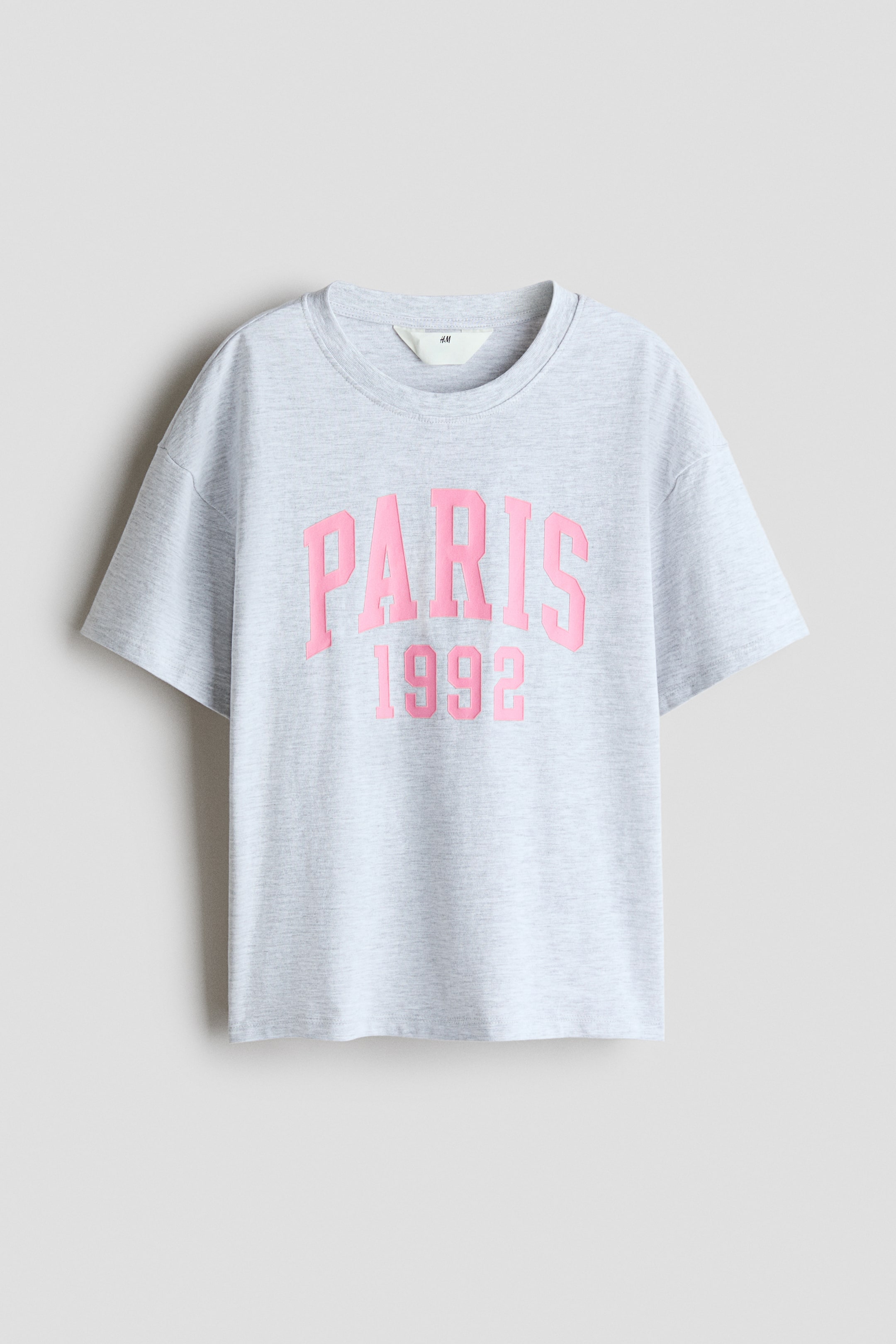 Größeres Bild ansehen: Ein hellgraues Baumwolljersey-T-Shirt in Melange-Optik zeigt seine Vorderseite mit einem pinkfarbenen "PARIS 1992" Grafikdruck. Es ist mit einem runden, gerippten Halsausschnitt und überschnittenen Schultern gestaltet.