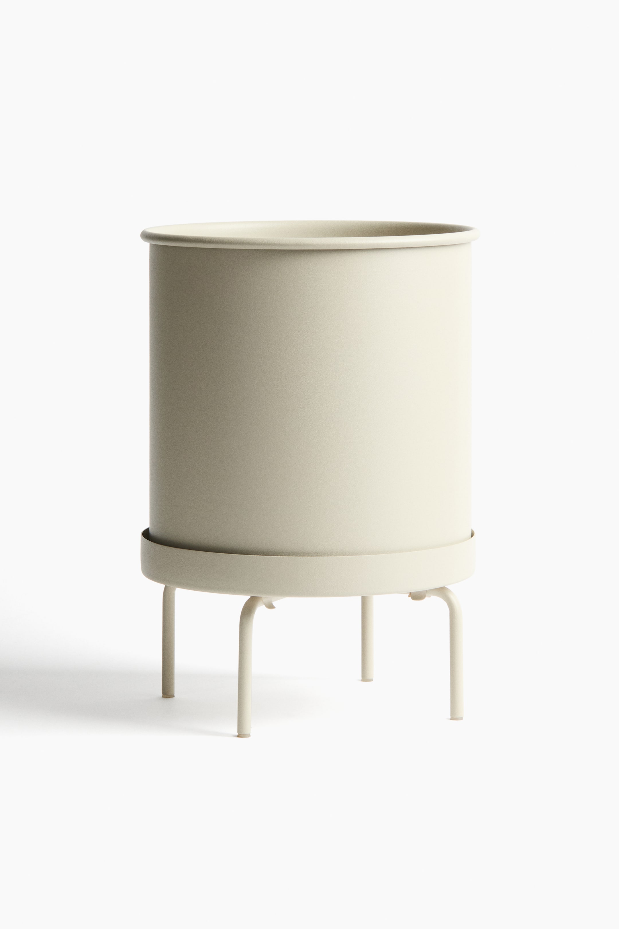 View larger image: A light beige cylindrical metal plant pot sits on a matching metal plant stand, shown from the front. The pot features a rolled rim, while the stand includes a circular saucer to hold the pot and four bent legs for support.