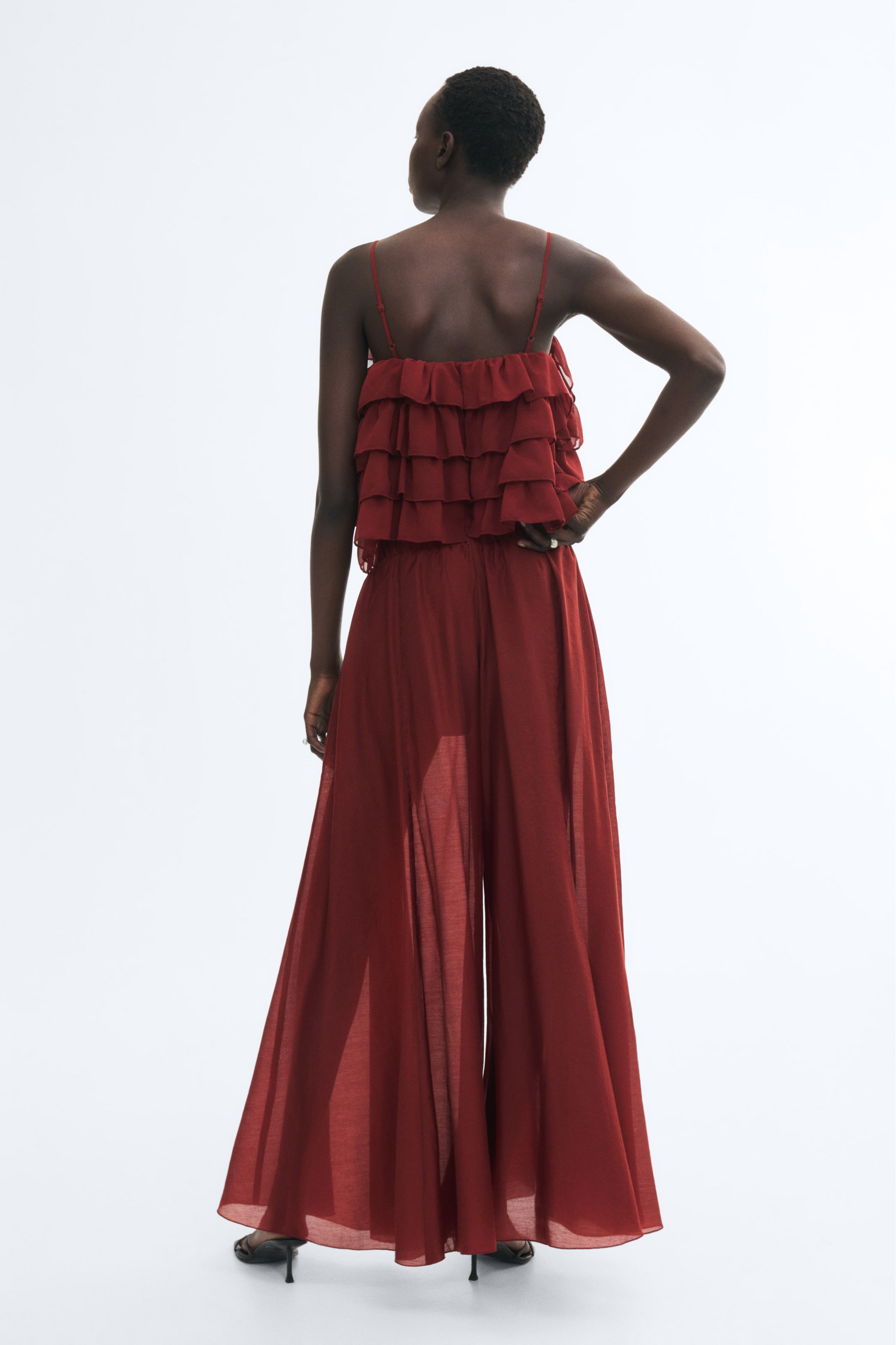 View larger image: Oversized, wide-leg pants in a sheer red fabric are worn by the model, who is standing with their back to the camera. The pants feature a flared silhouette with a shorter lining visible underneath and are styled with a matching ruffled camisole top.