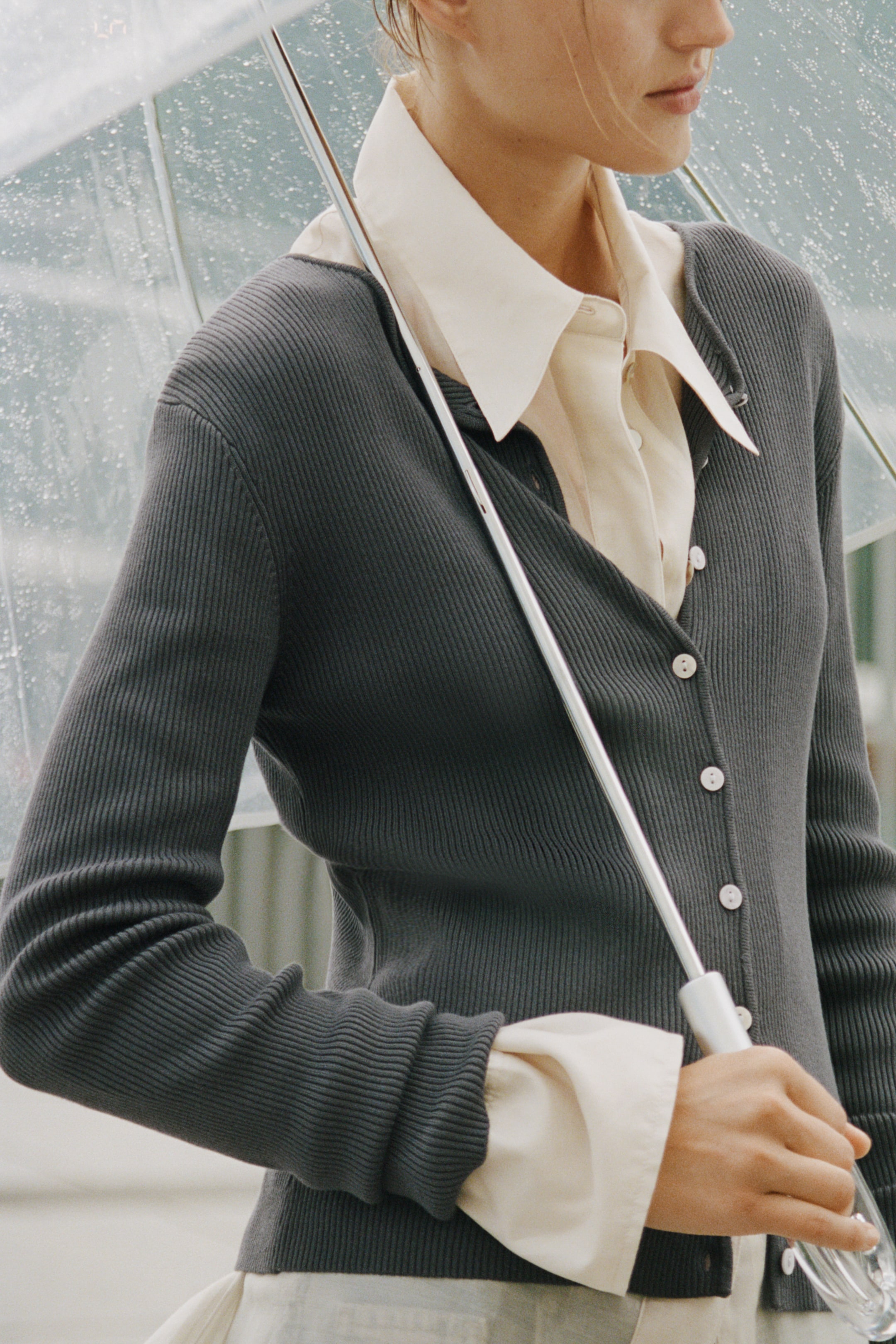 Ver imagem maior: Um cardigan canelado cinzento-escuro está a ser usado pela modelo sobre uma camisa creme, com colarinho e botões à frente. O cardigan apresenta um decote em V, botões de cor clara na frente e mangas compridas, enquanto a modelo segura um guarda-chuva transparente.