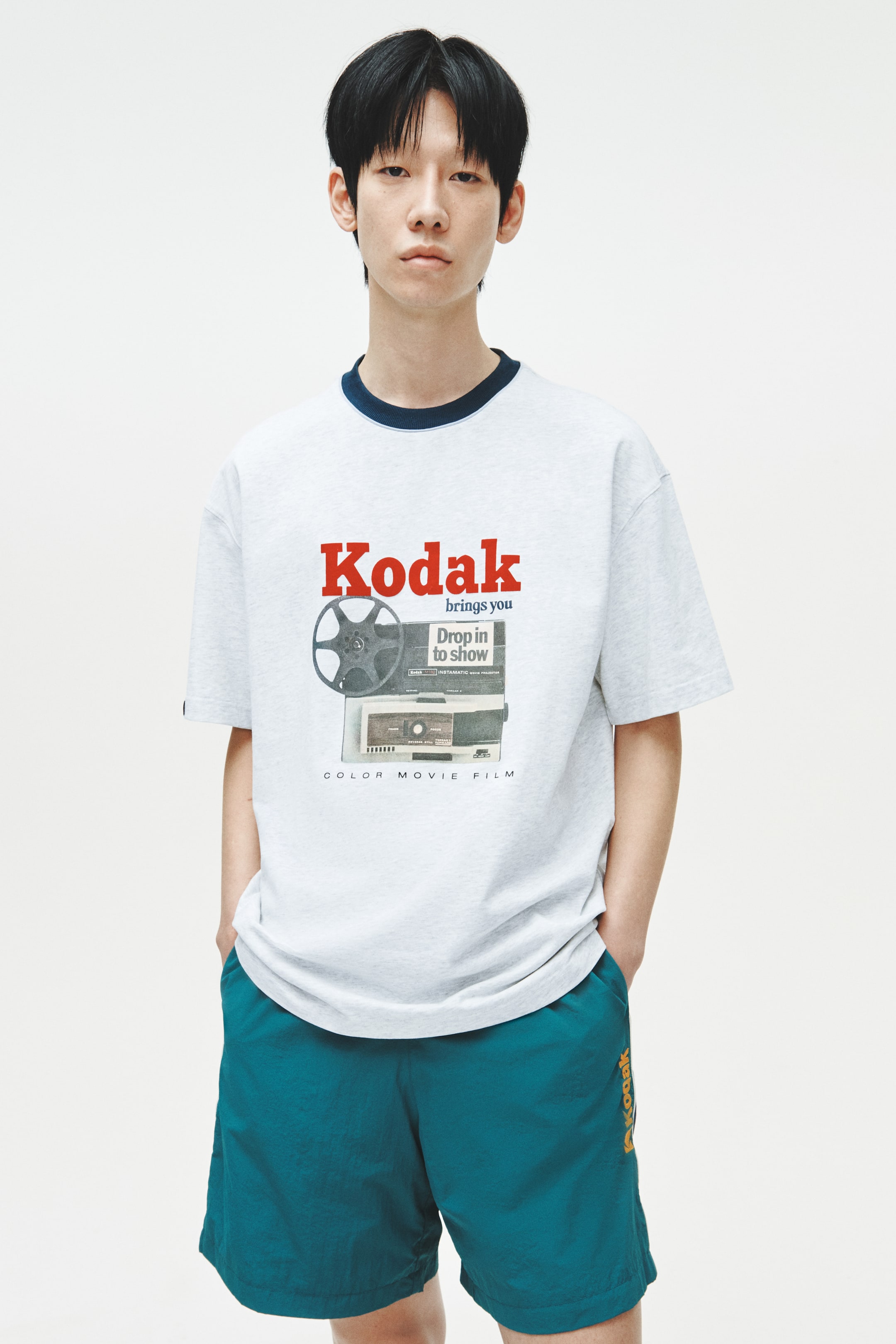 View larger image: A light grey t-shirt with a navy blue ribbed crew neck and short sleeves showcases a vintage-style graphic on the front, featuring the red "Kodak" logo, accompanying text, and an image of a film reel and projector. Complementing this are teal shorts with an elastic waistband, side pockets, and an orange "Kodak" logo printed vertically on the right leg.