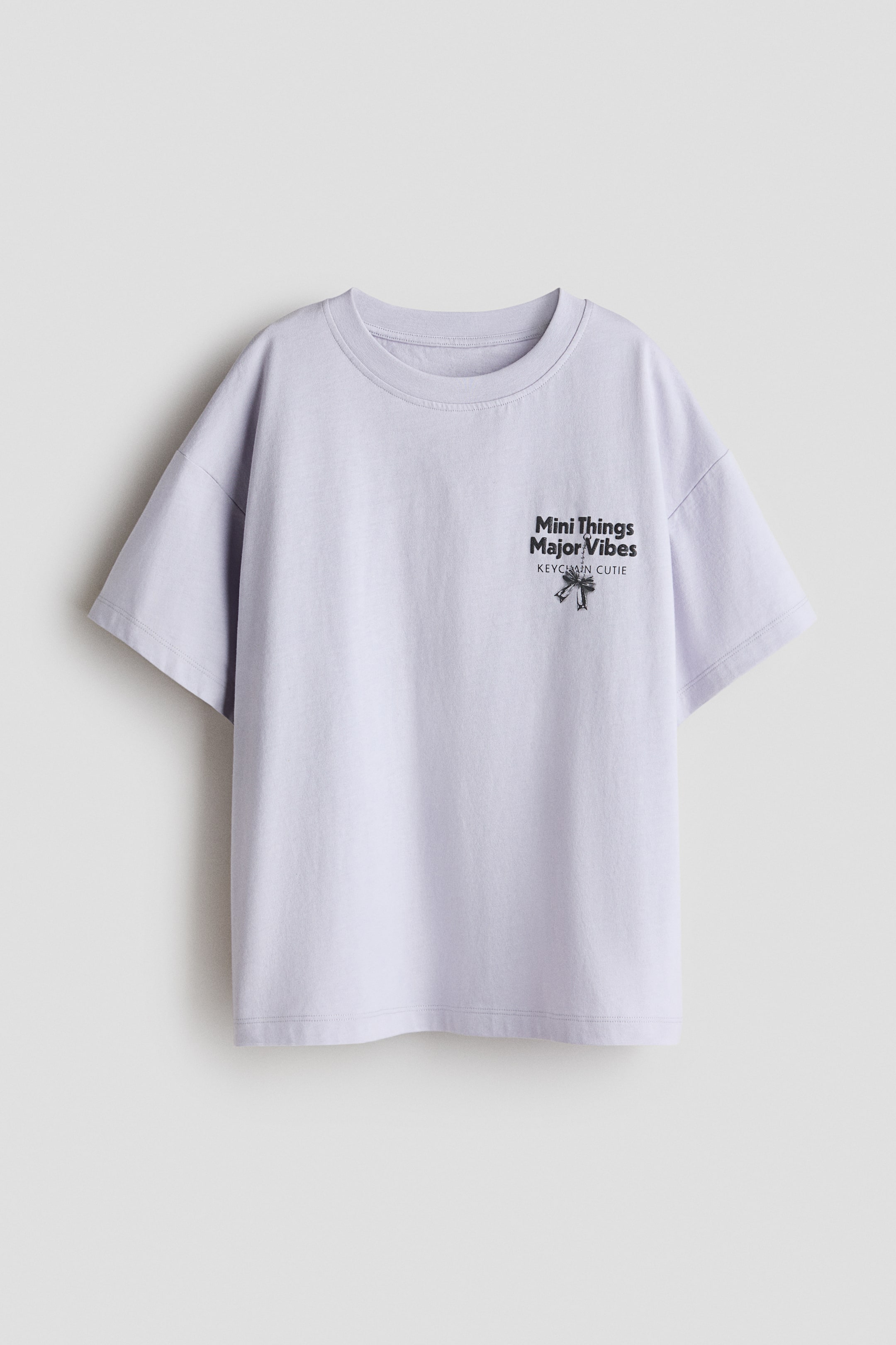 Προβολή μεγαλύτερης εικόνας: Μπροστινή όψη ενός υπερμεγέθους ανοιχτού μοβ μπλουζιού από μαλακό, τυπωμένο βαμβακερό ζέρσεϊ, που δείχνει στρογγυλή, ριμπ-κομμένη λαιμόκοψη, χαμηλωμένους ώμους και ίσιο τελείωμα. Ένα μαύρο γραφικό σχέδιο εμφανίζεται στο αριστερό στήθος, με το κείμενο 'Mini Things Major Vibes' και 'KEYCHAIN CUTIE' με έναν μικρό σχεδιασμό γοητείας.