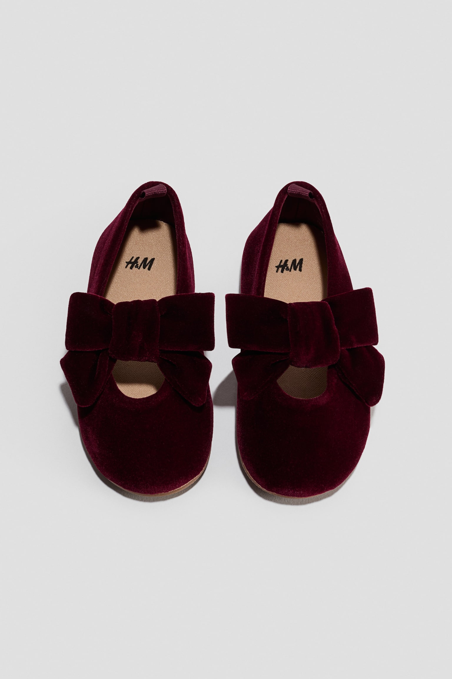 Een paar bordeauxrode Mary Jane-schoenen van fluweel wordt van bovenaf en van voren afgebeeld, met een ronde neus en een grote, decoratieve fluwelen strikband over de wreef. De schoenen tonen een lichtbeige binnenzool met een gedrukt logo en een klein treklusje aan de hiel.
