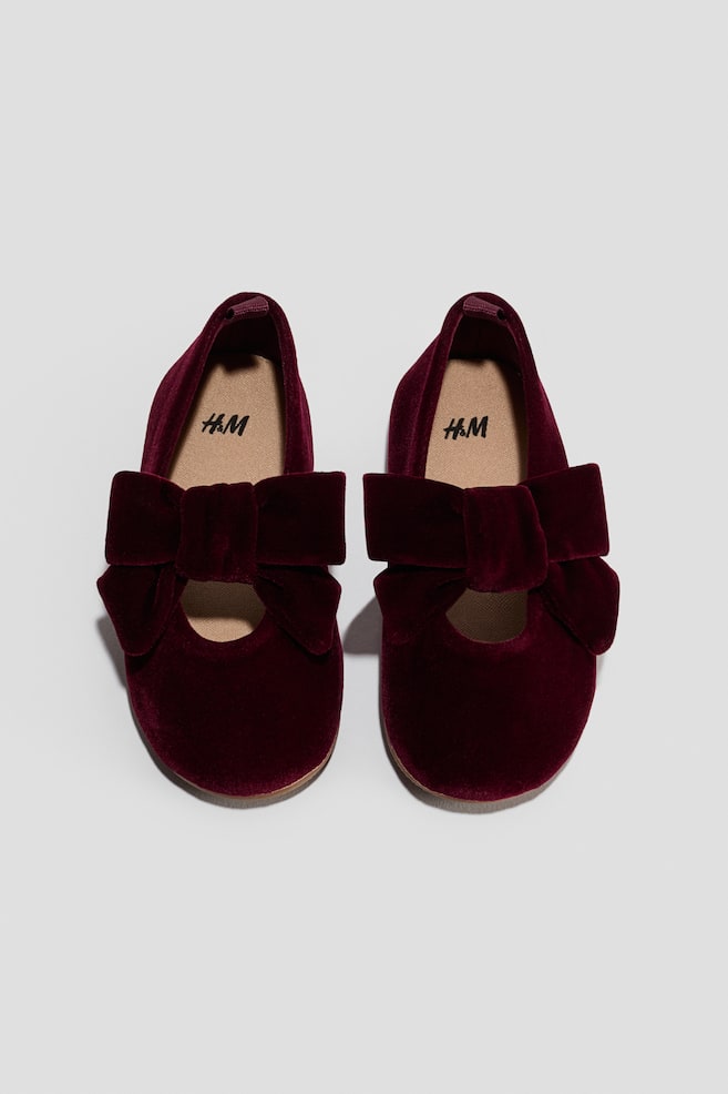 A pair of burgundy velvet Mary Jane shoes is shown from a top-down angle facing front, featuring a rounded toe and a large, decorative velvet bow strap across the instep. The shoes are displaying a light beige insole with a printed logo and a small pull tab at the heel. - 1