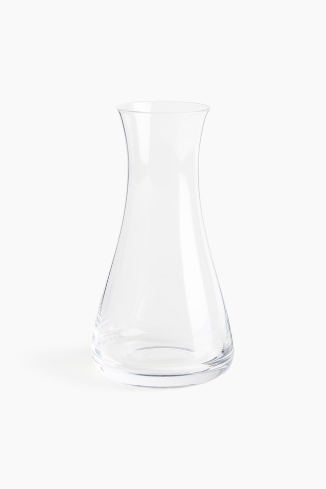 A small clear glass carafe is standing facing the front, featuring a wide, conical base that tapers upwards into a narrow neck and ends with a gently flared opening. - 1