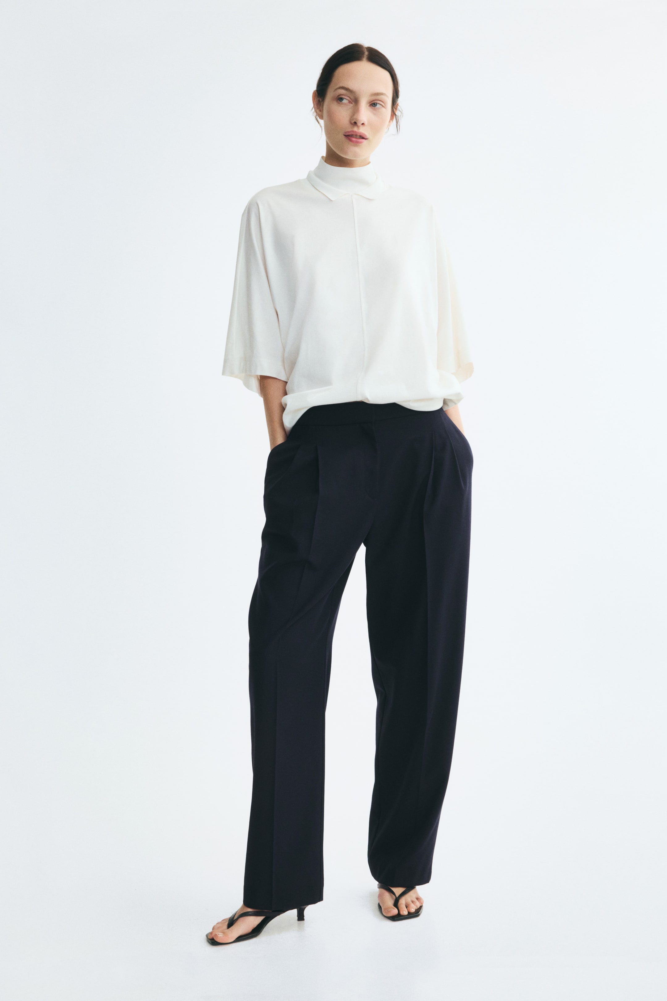 View larger image: Tailored, loose-fit pants in dark navy are worn by the model, who is standing with her hands resting in the side pockets. The high-waisted pants feature front pleats and creases down the barrel legs, styled with a tucked-in white short-sleeve top and black heeled sandals.