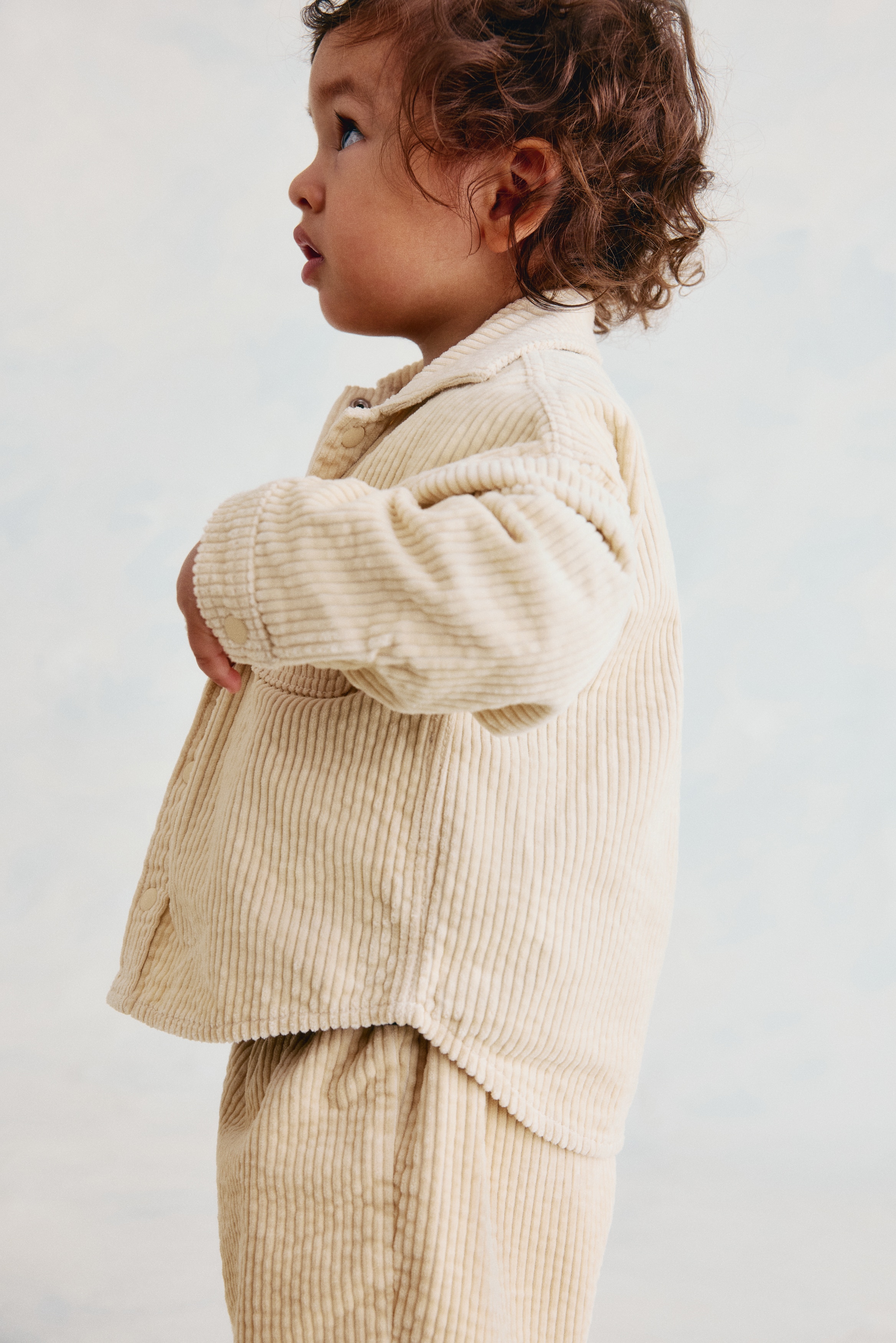A cream corduroy shirt is shown from the front, featuring a pointed collar and a full button-front placket with round, matching buttons. It includes a single patch pocket on the left chest, long sleeves, and a curved hem.
